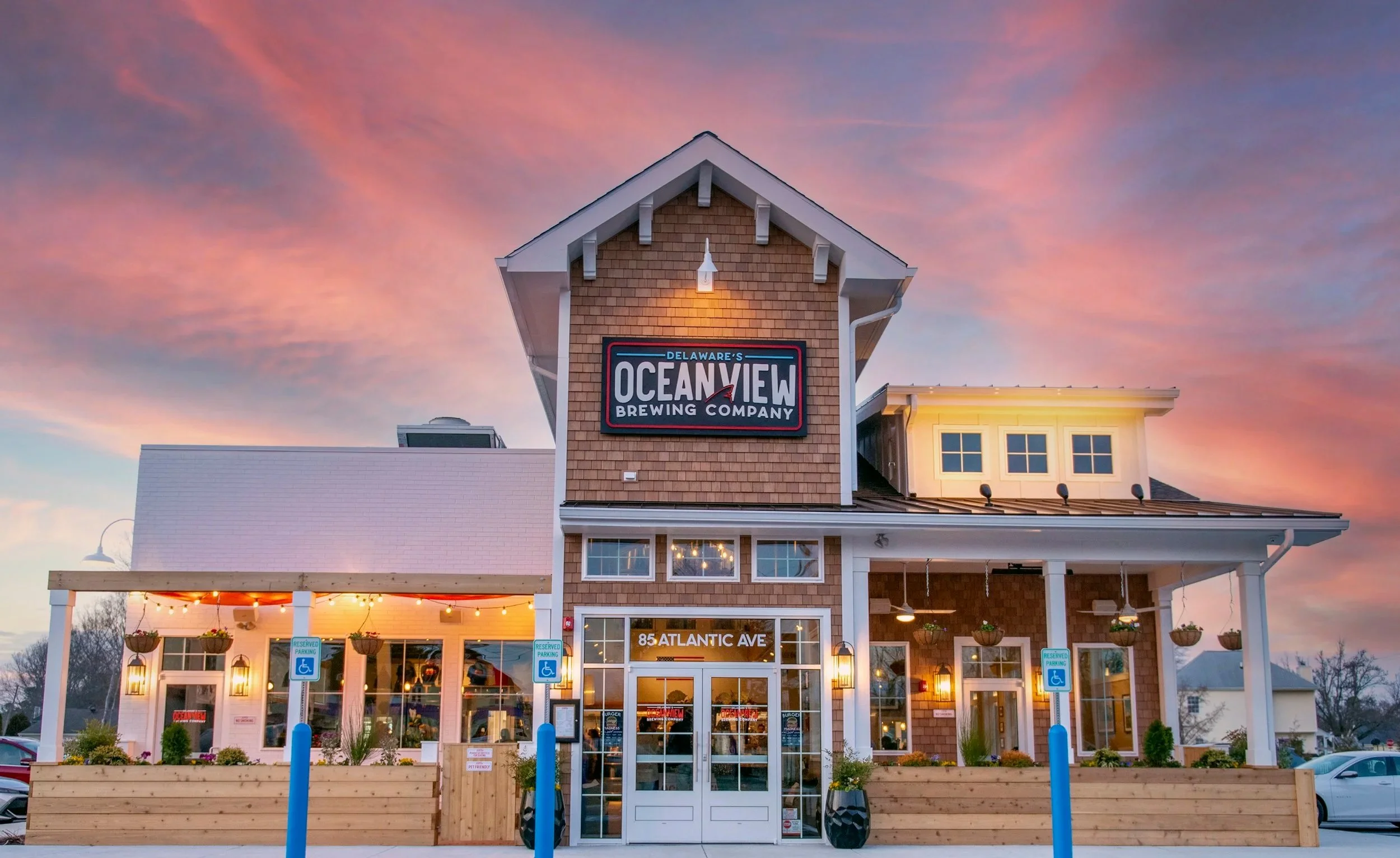 A restaurant named Oceanview Brewing Company located at 85 Atlantic Ave, with a sunset sky in the background. The building has a brick and white exterior with a sign displaying the restaurant's name. There are outdoor seating and reserved parking signs in front.