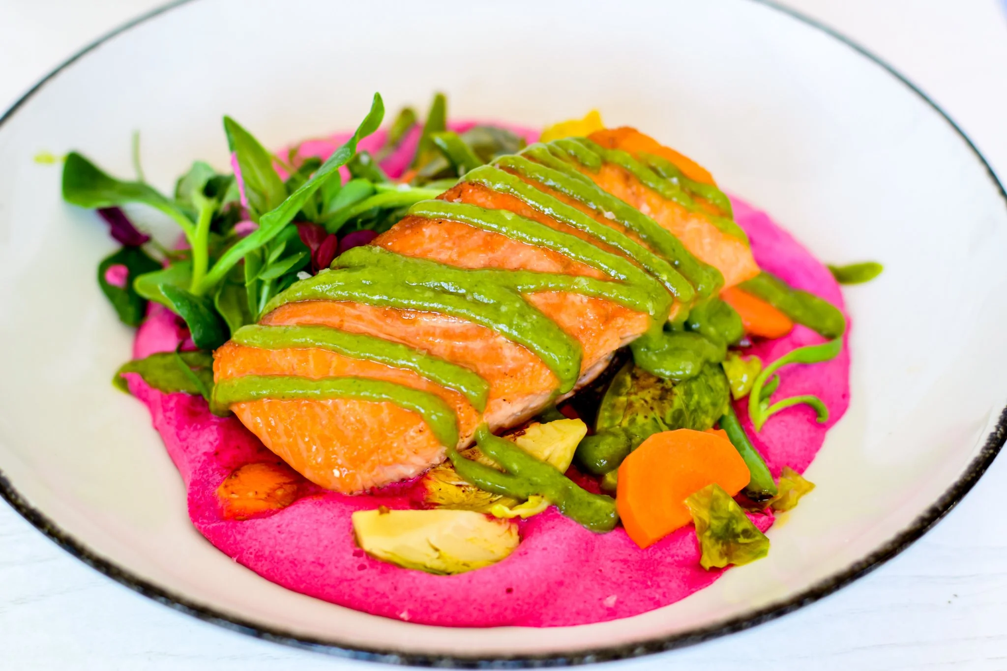 A dish featuring a cooked piece of salmon topped with green sauce, served on a bed of colorful vegetables and pink puree in a white bowl with a black rim.