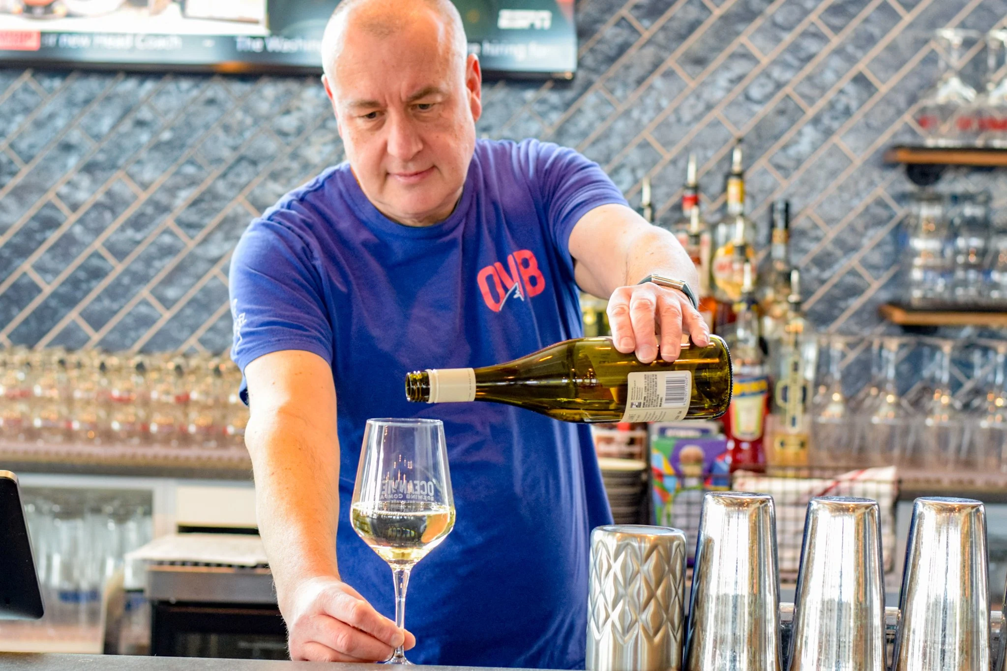 A man in a blue shirt pouring white wine into a wine glass in a bar or restaurant setting.