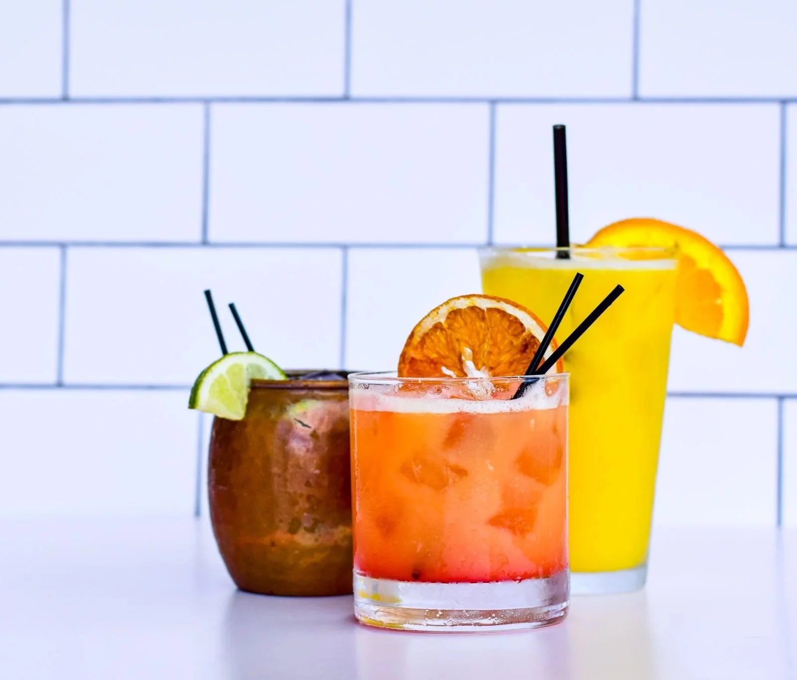 Three colorful cocktails with lime, orange, and lemon garnishes on a white surface against a white tiled wall.