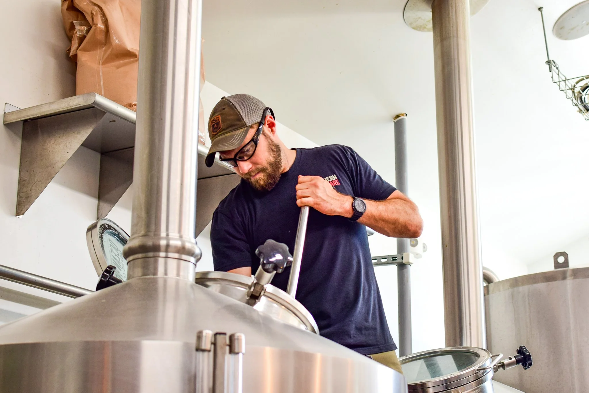 A man wearing a baseball cap, glasses, and a black t-shirt working on a large stainless steel brewing or processing vessel in a laboratory or industrial setting.