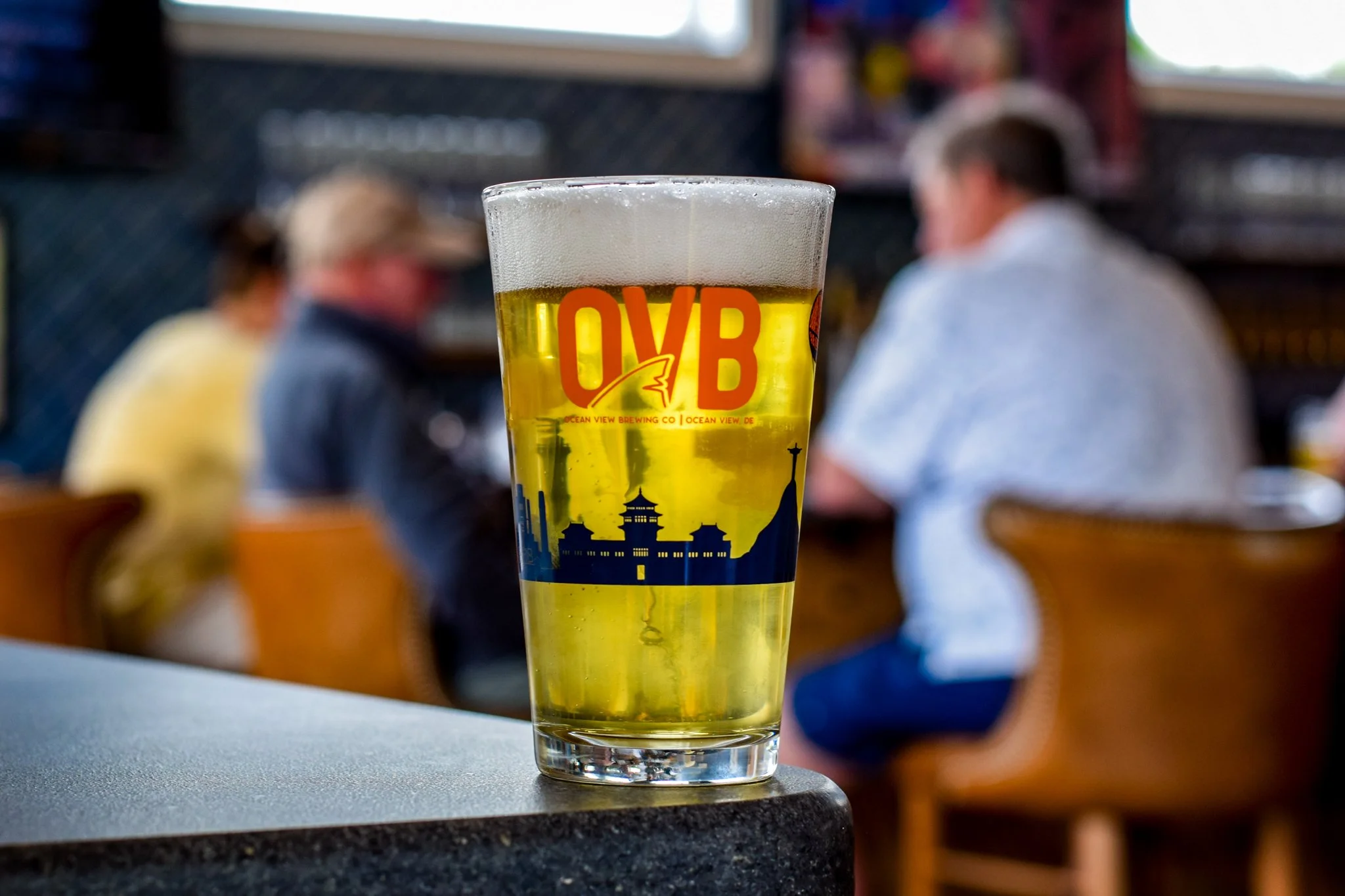 A pint glass filled with beer sitting on a table at a bar or restaurant, with a blurred background of two people sitting at a table and a window.