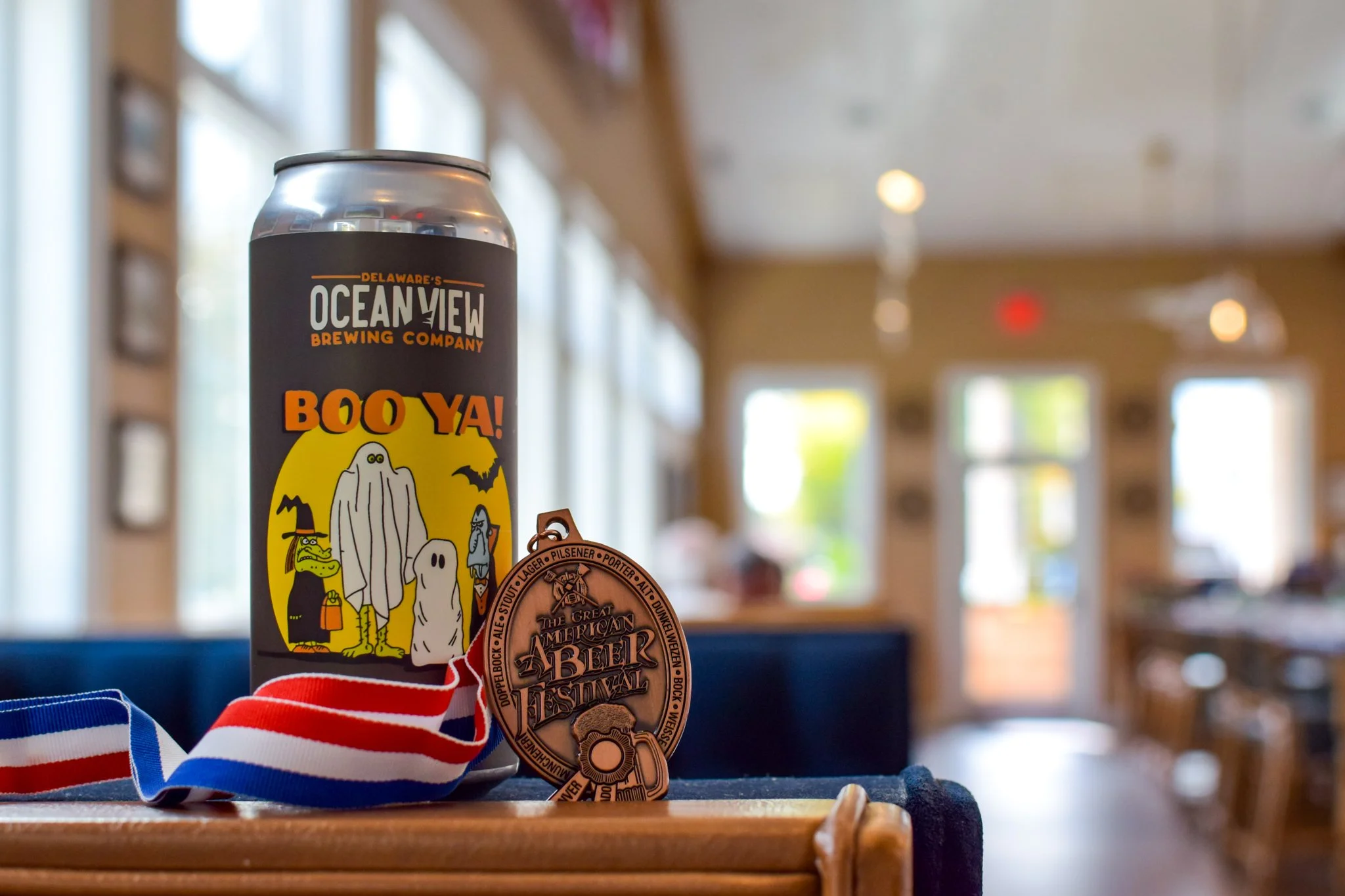 A can of Delaware's Ocean View Brewing Company's Boo Ya Halloween-themed beer, accompanied by a medal or tag from a beer festival, and a red, white and blue striped ribbon, all on a wooden table inside a room with large windows and warm lighting.