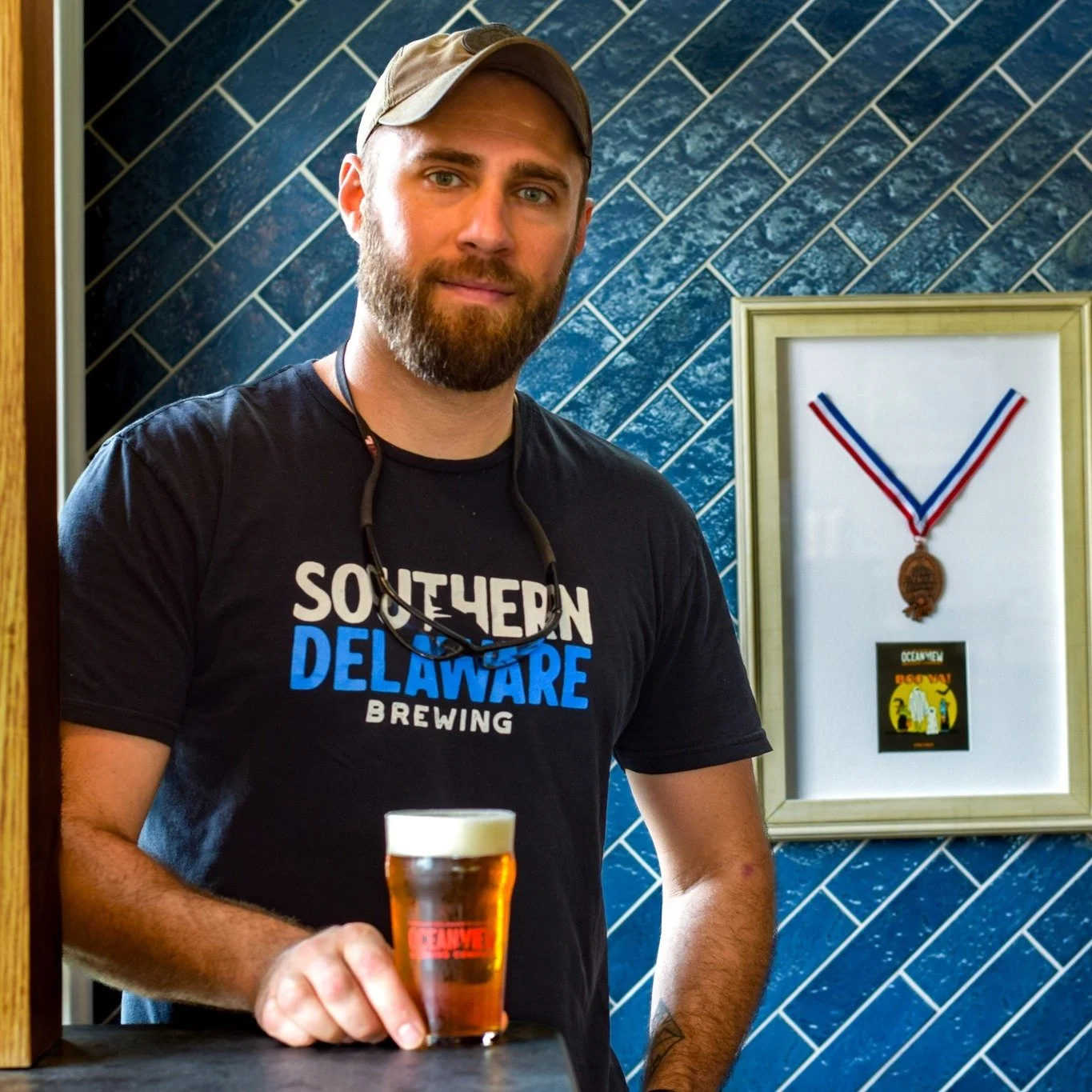 A man with a beard and wearing a cap, a black T-shirt with words "Southern Delaware Brewing" and sunglasses around his neck, holding a glass of beer, standing in front of a blue tiled wall with a framed medal and logo on it.