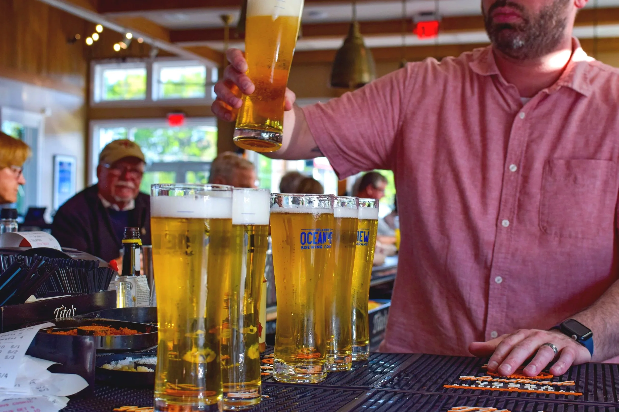 Four tall glasses of beer on a bar counter with a bartender pouring a beer in the background. The bar has a rustic, cozy interior with large windows and warm lighting.