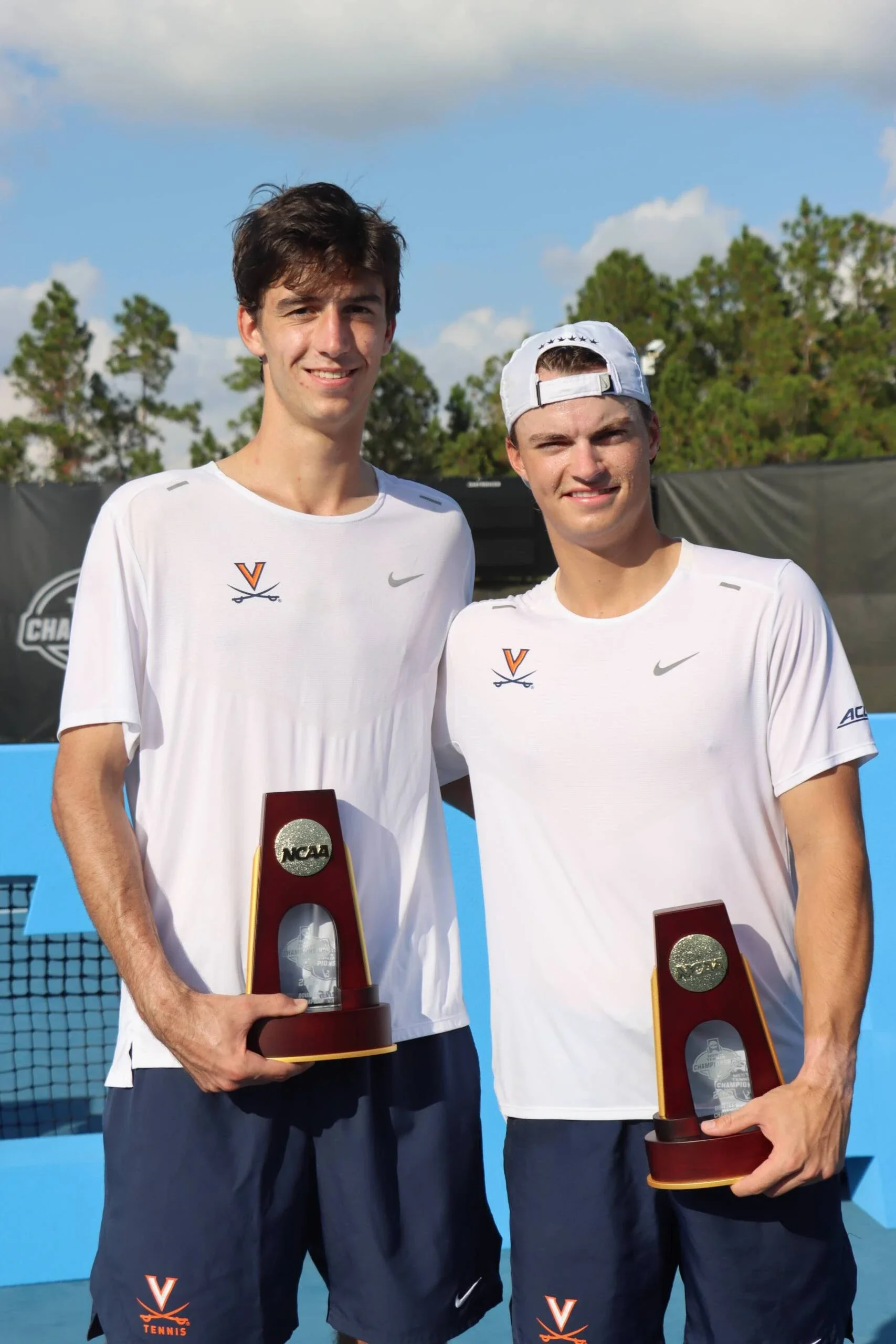 Dylan Dietrich, Zwei junge Männer in weißen Sportshirts und Shorts, die jeweils einen Pokal halten, stehen auf einem Tennisplatz. Im Hintergrund sind Bäume und ein blauer Himmel.