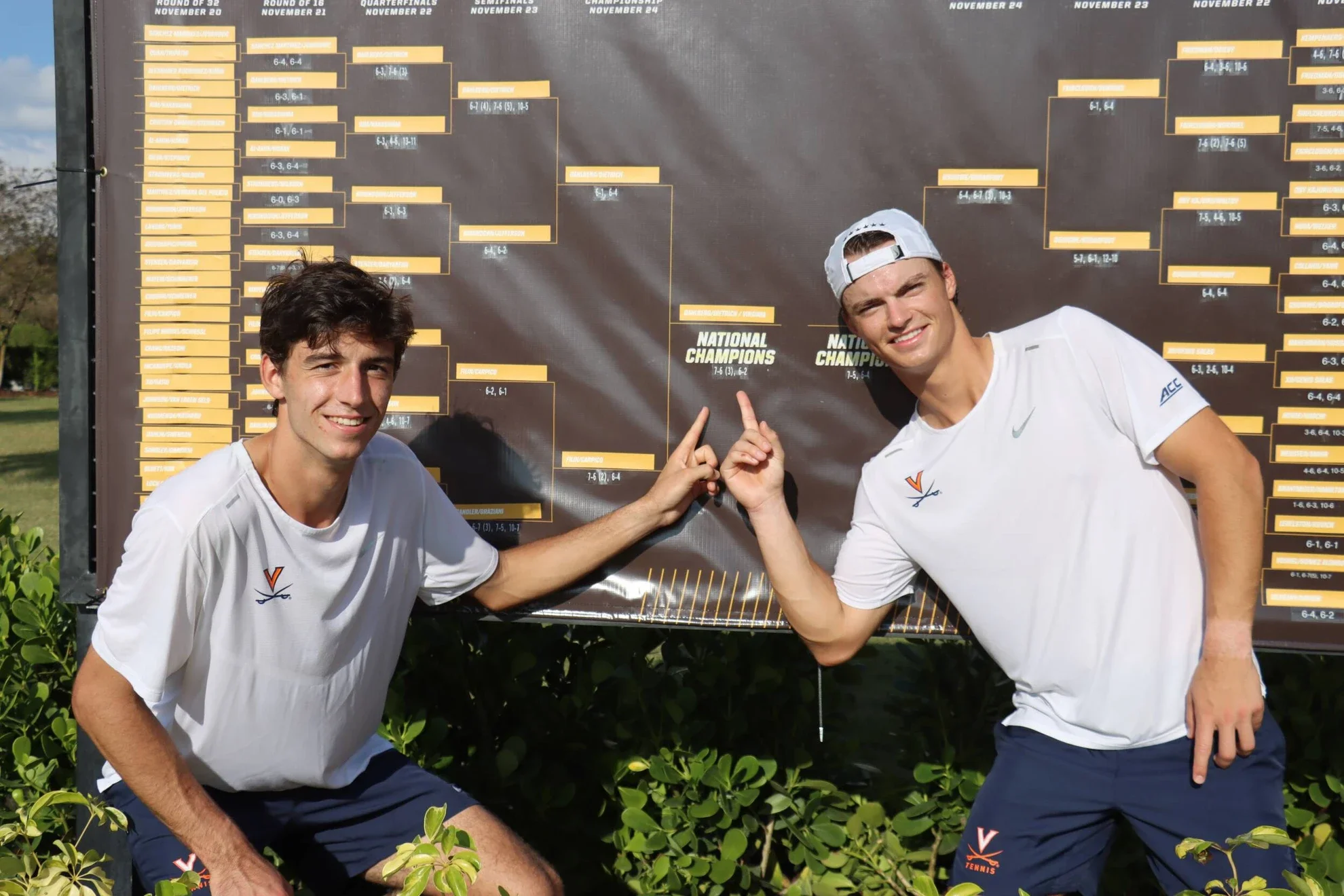 Zwei junge Männer in weißen Tennisshirts und blauen Shorts stehen vor einem Turnierbaum und zeigen auf die Schilder, die die Meisterschaftsstände anzeigen, im Freien mit Grünflächen.