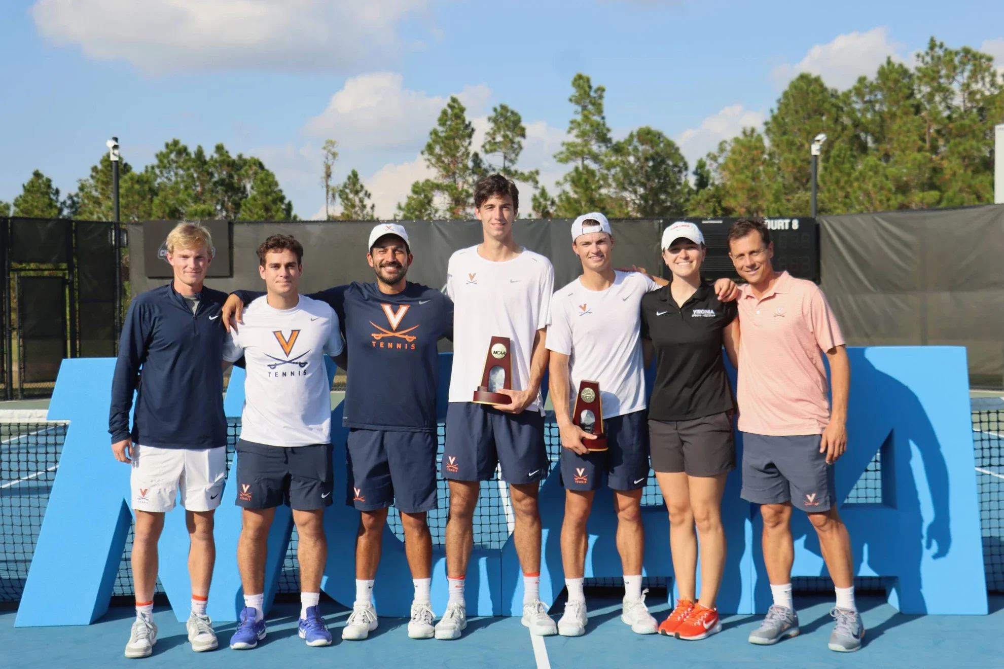 Sieben Tennisspieler stehen auf dem Tennisplatz nach einem Turnier, einige halten Pokale. Es ist sonnig, im Hintergrund Bäume und ein blauer Himmel.