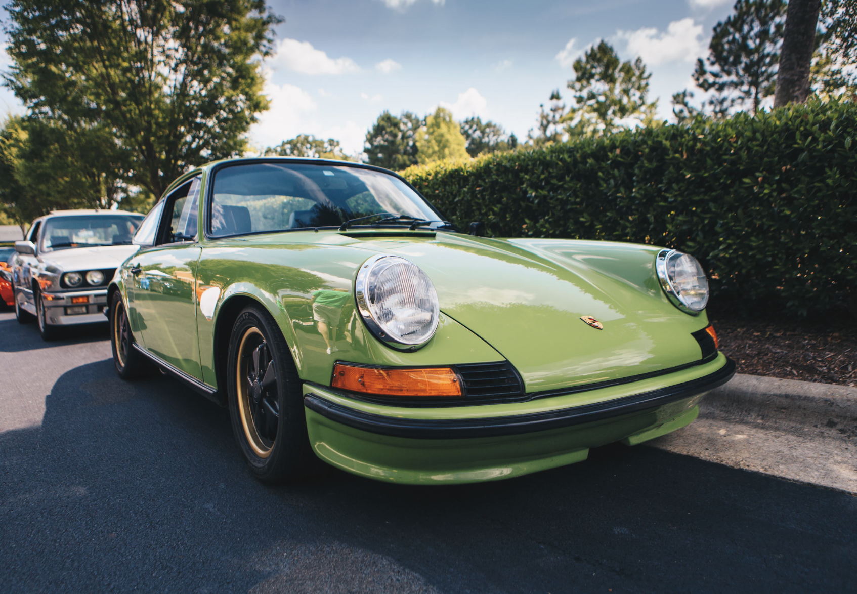 A green vintage sports car parked on a street, with a silver car behind it, surrounded by trees and bushes.