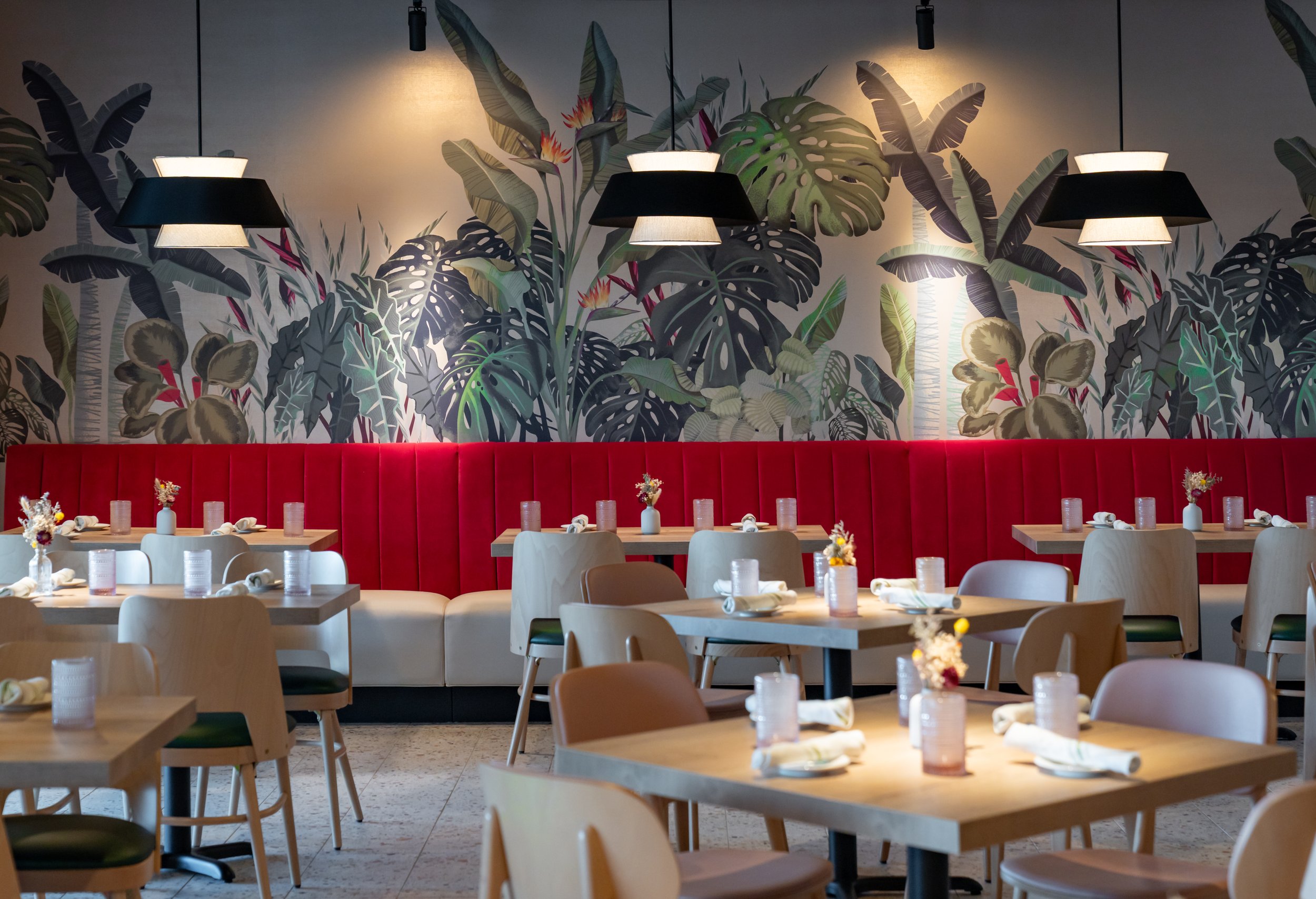 A modern restaurant interior with a tropical leaf mural on the wall, a red banquette seating area, and light-colored wooden tables and chairs set with napkins and small vases of flowers.