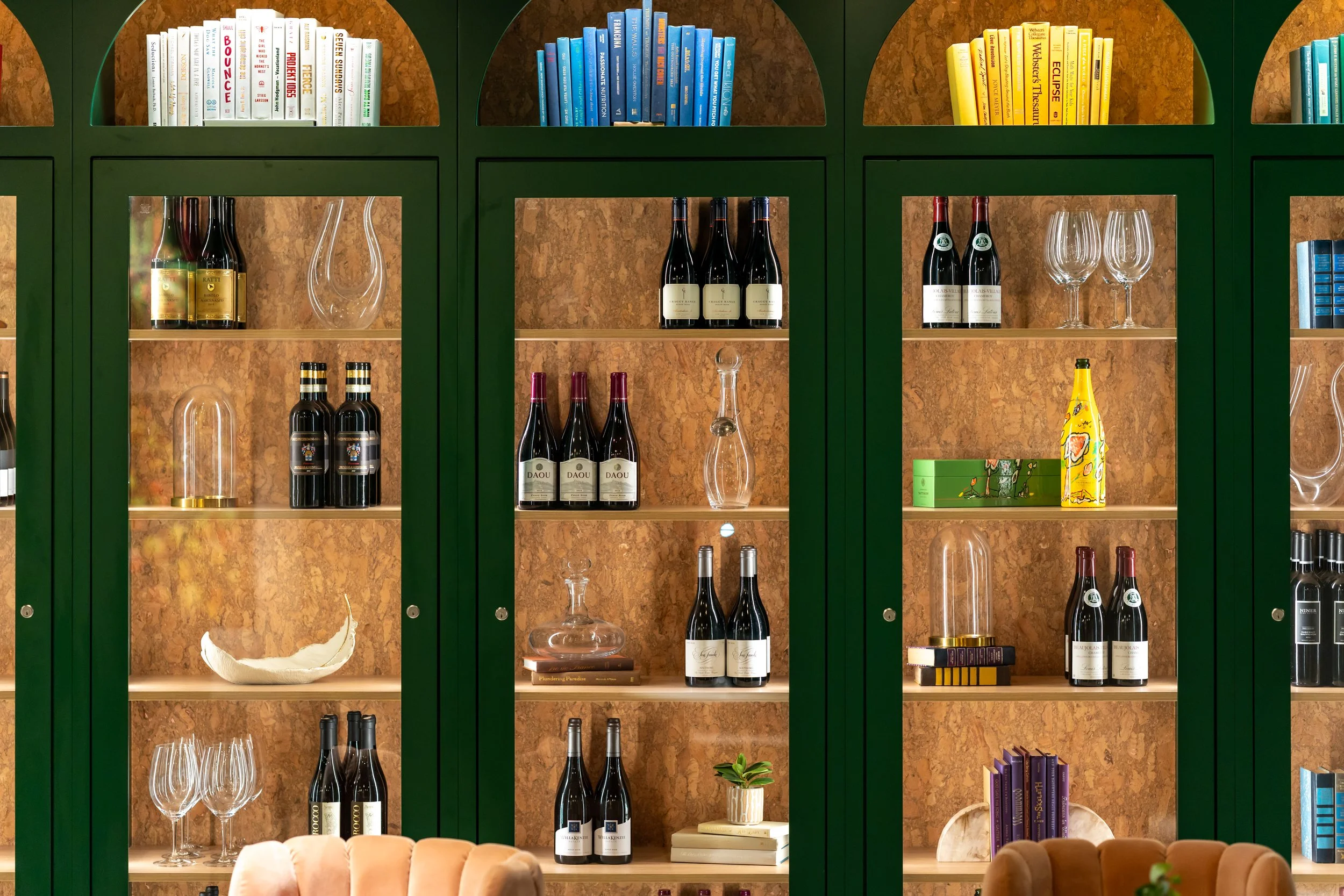 Green shelving unit with wine bottles, glasses, decorative objects, and books against a wood-paneled background.