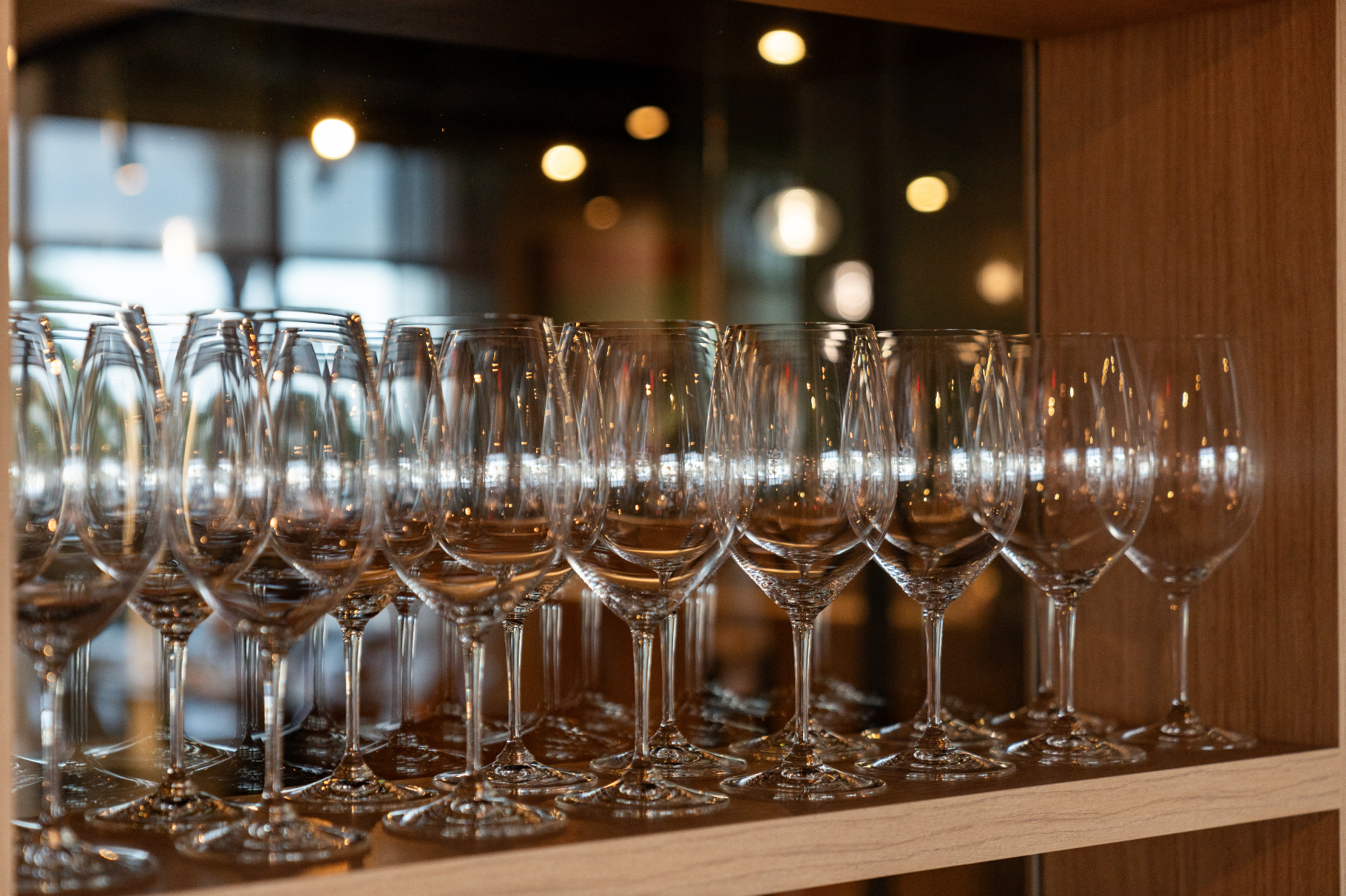 Multiple empty wine glasses arranged on a wooden shelf, with a blurred indoor background and warm lighting.