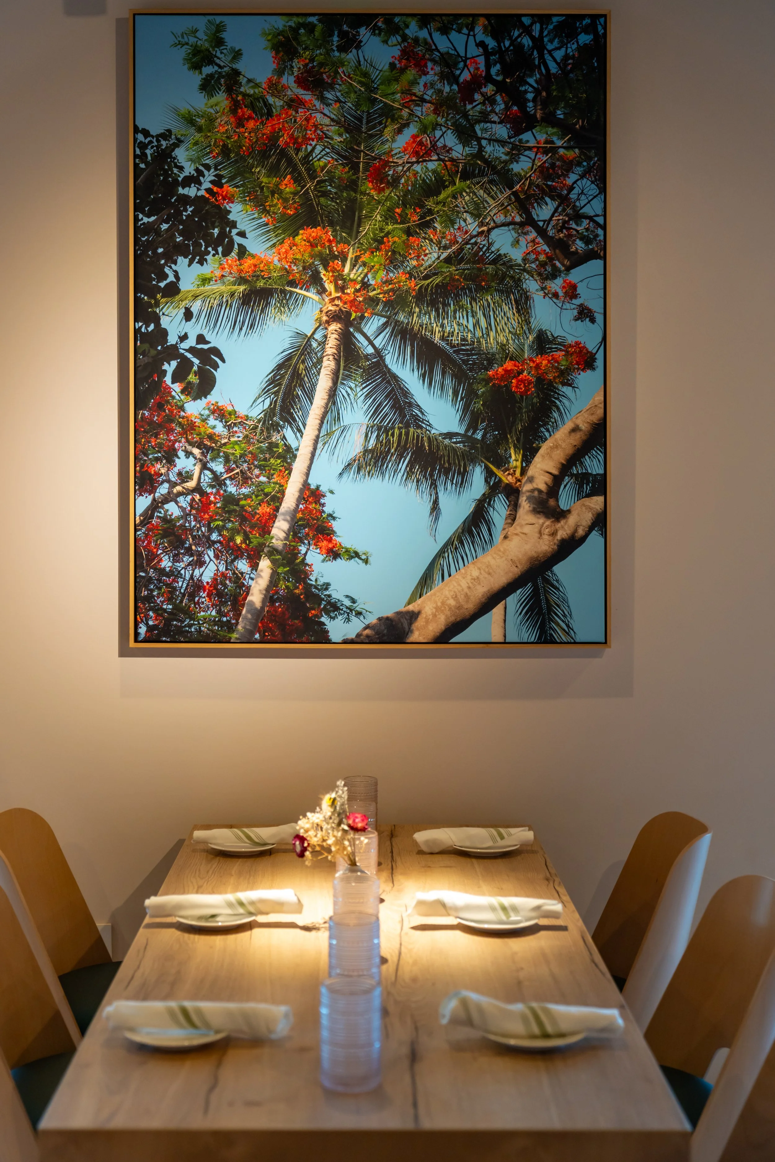 A dining table set for four with white plates, napkins, and glasses in front of a large framed photograph of palm trees and a blue sky.