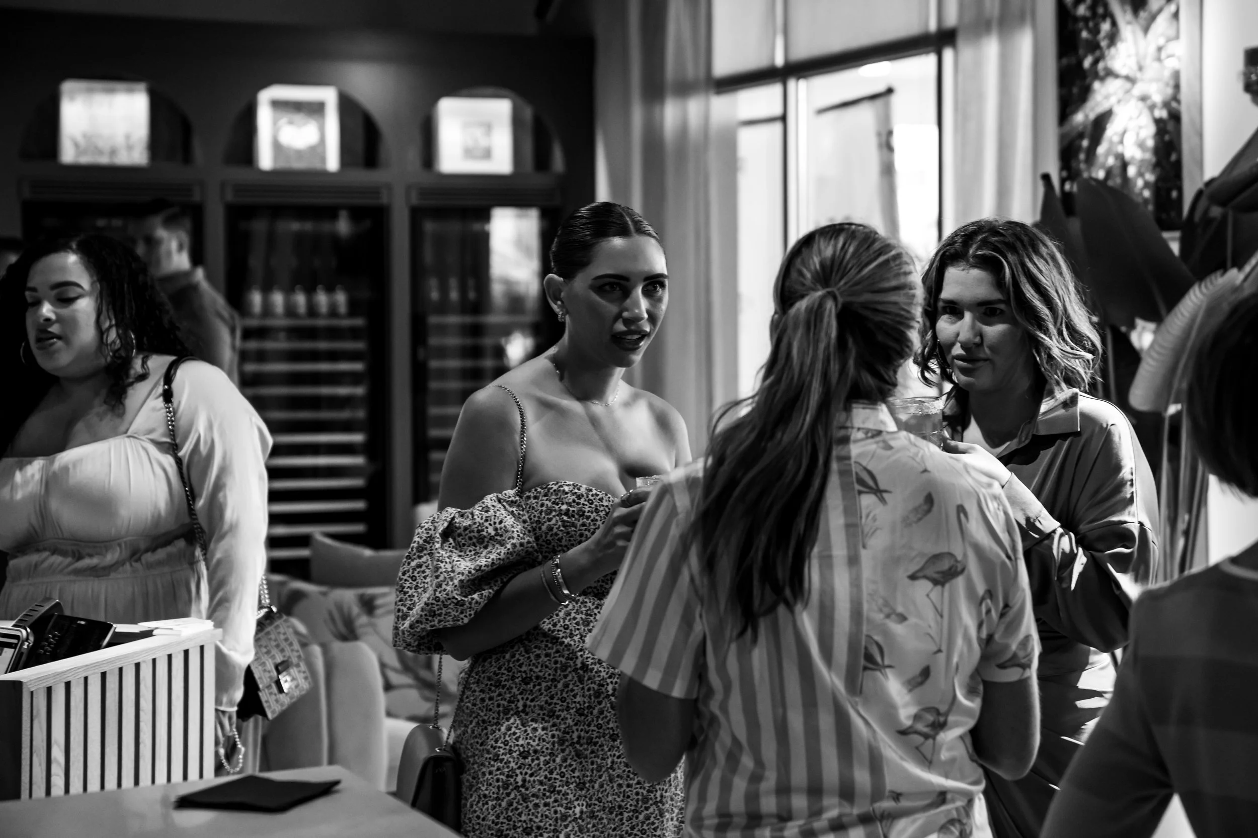 Group of women socializing in a well-lit indoor space, one woman holding a drink and talking to others, some wearing patterned clothing.