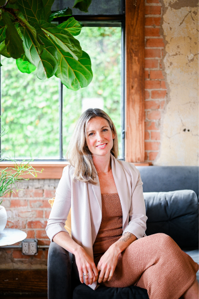 Photo of Jaime Bell sitting on a couch smiling at the camera. She is wearing a brown dress with a light brown jacket and there is a plant behind her.