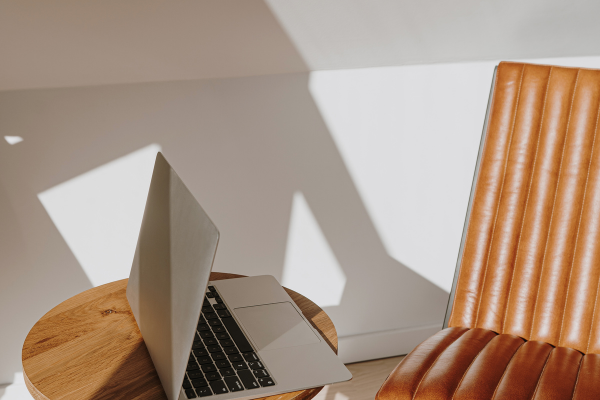 Open laptop on a round wooden table next to a brown leather chair in a bright room with sunlight and shadows on the wall.