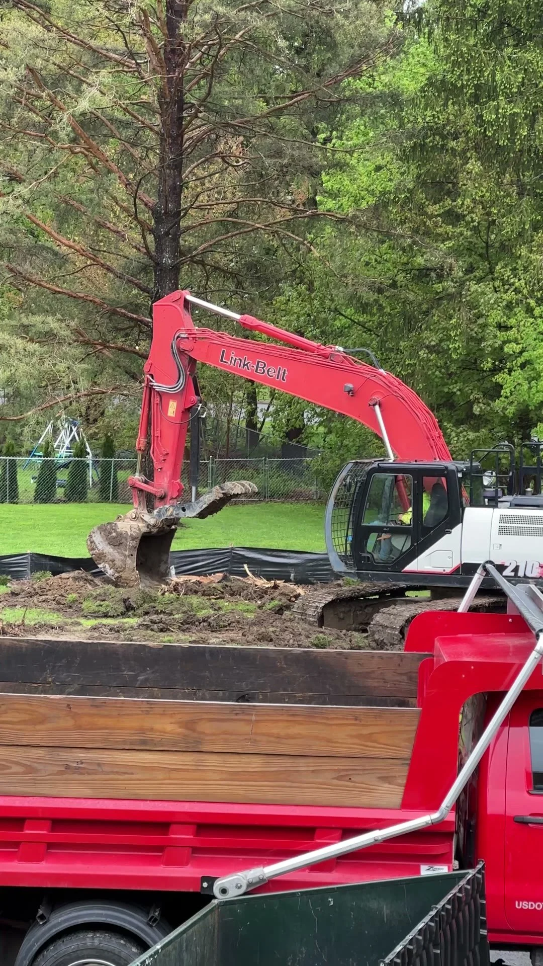 A red excavator with the label "Link-Belt" is digging a hole in a grassy yard, pulling up dirt and soil. The background shows trees and a fence.