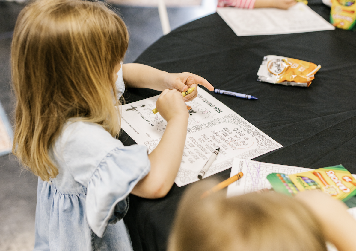 A young girl with red hair coloring a festive worksheet at a table, with crayons, markers, snacks, and other coloring pages nearby.