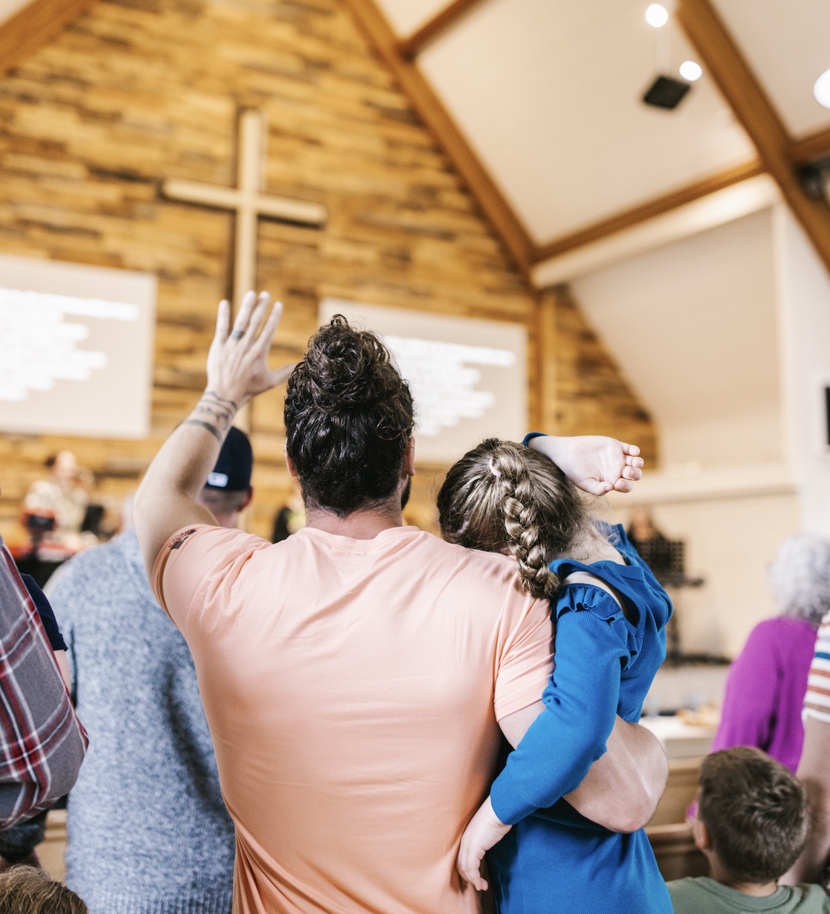 A man holding a young girl during a worship service in a church with a cross on the wooden wall at the front.