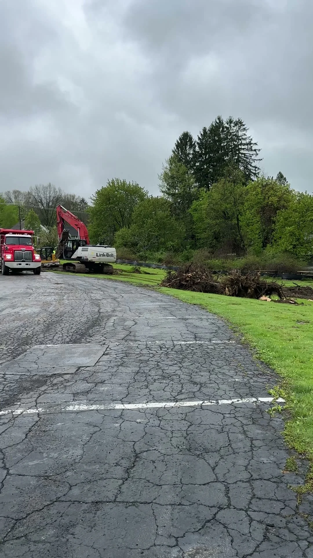 A parking lot with cracked asphalt, showing fallen tree branches and construction equipment, including a red excavator and a white Link-Belt machine, near a green grassy area with trees under a cloudy sky.