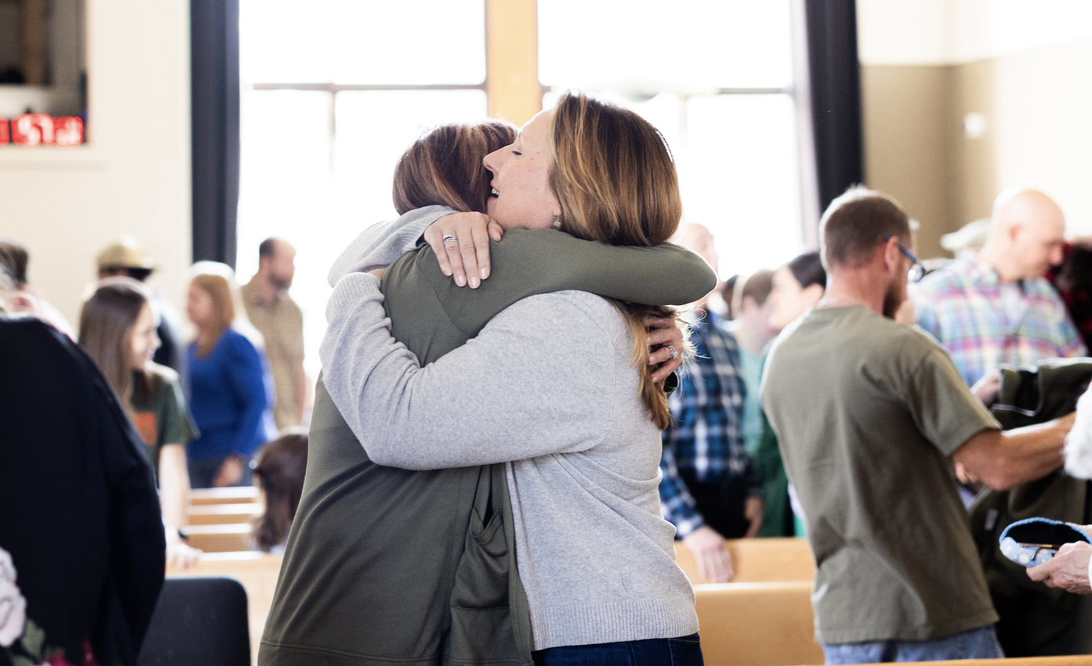Two women hugging lovingly in a crowded room, with people in the background engaging in conversation.