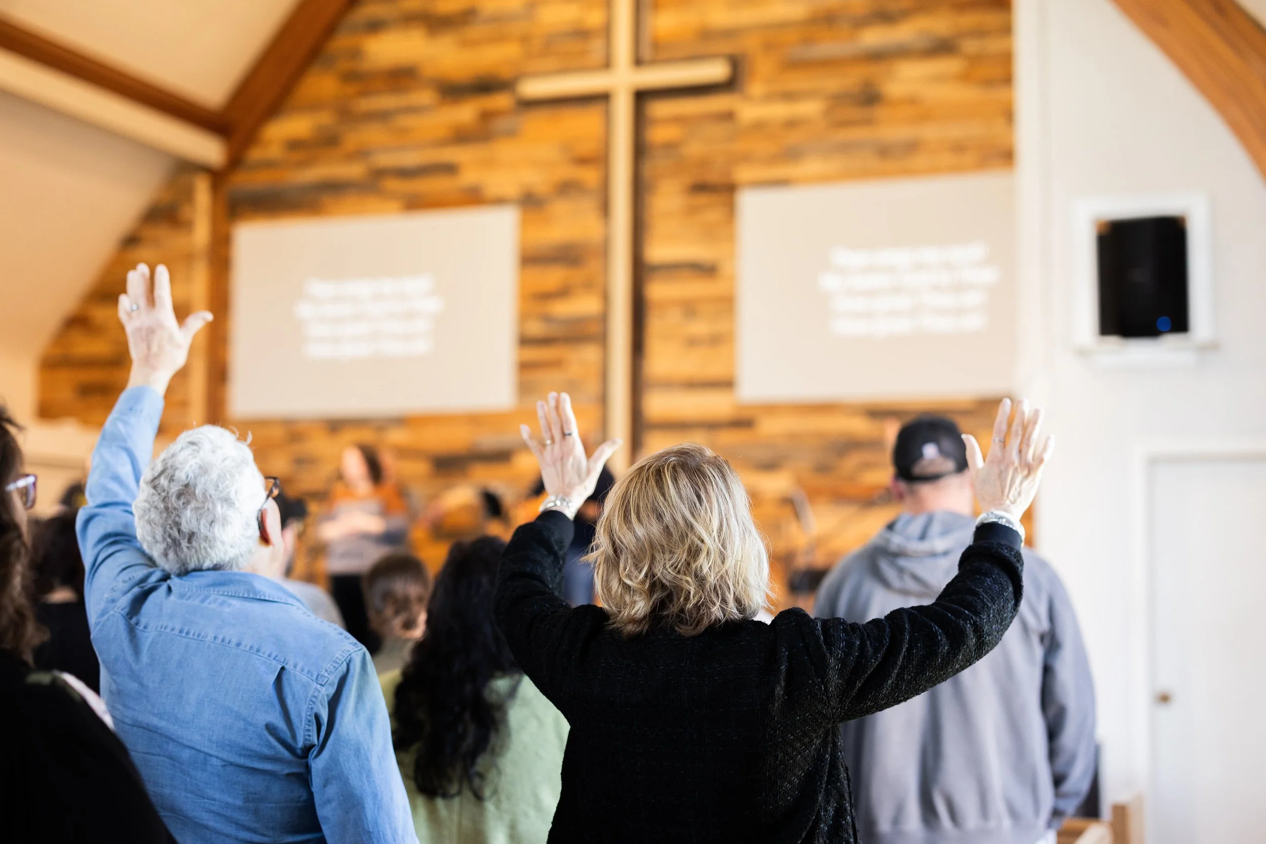 People attending a church service, raising their hands during a prayer or worship in a church with wooden walls and a cross on the altar.