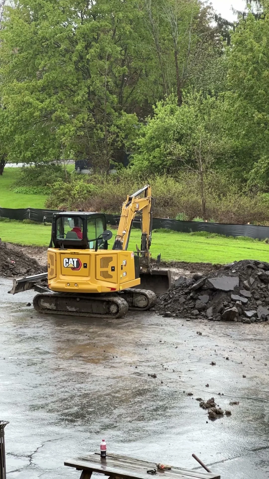 Construction site with excavator moving dirt on a cloudy day, trees in the background, a Coke bottle and tools on a bench in the foreground.