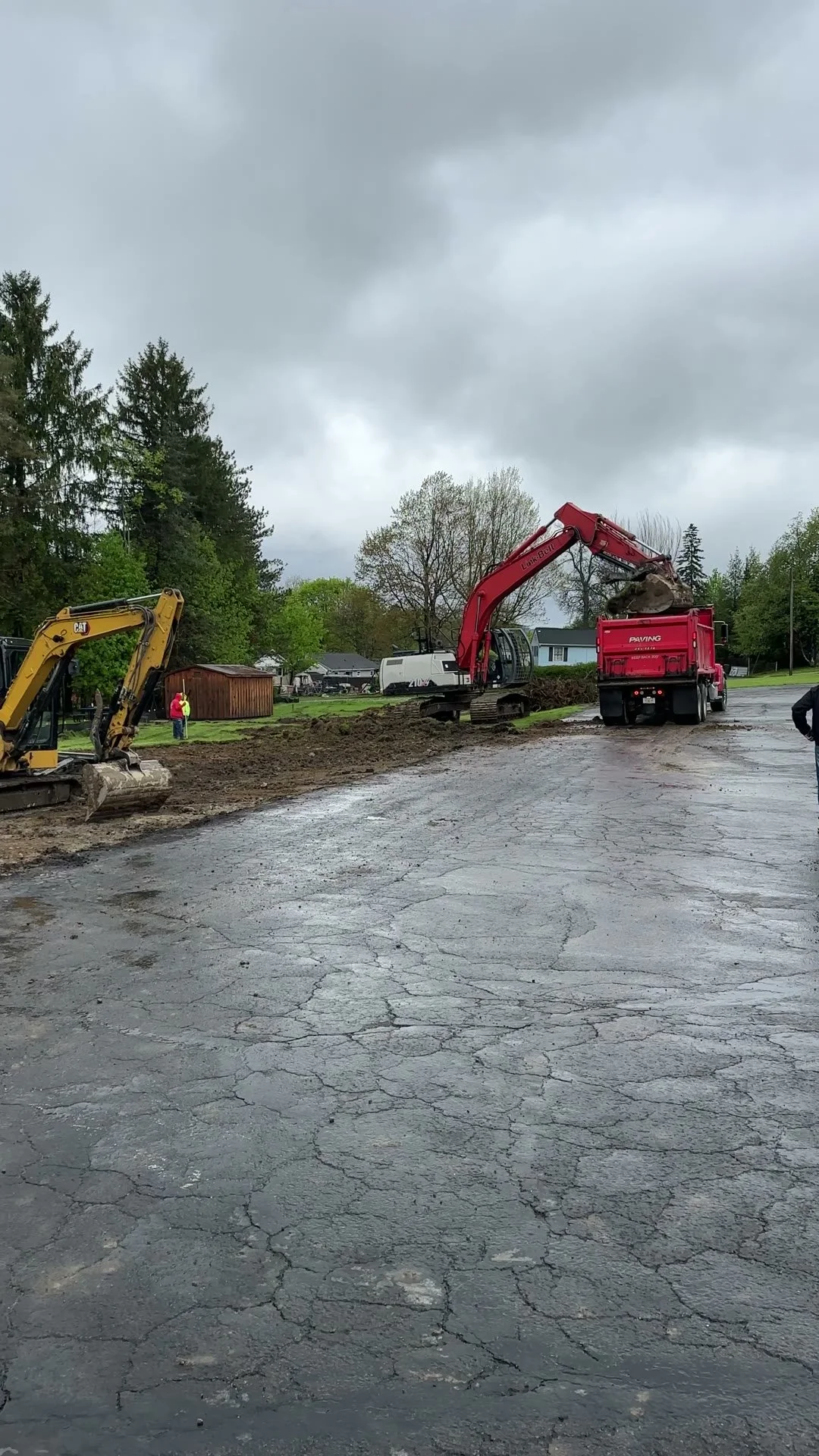 Construction workers and heavy machinery, including a yellow excavator and a red excavator, are working on road repairs in a residential area on a cloudy day.