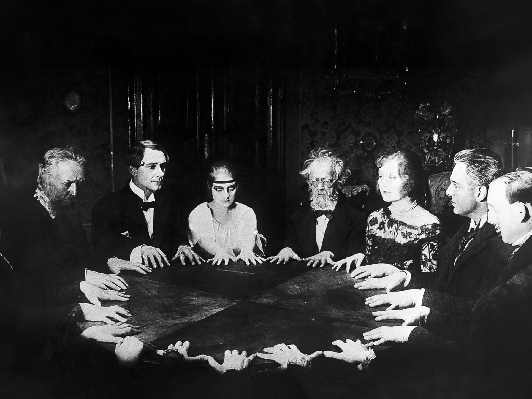 Black and white film scene of nine people with their hands on a large square table, all looking down or to the side, with a dark ornate background.