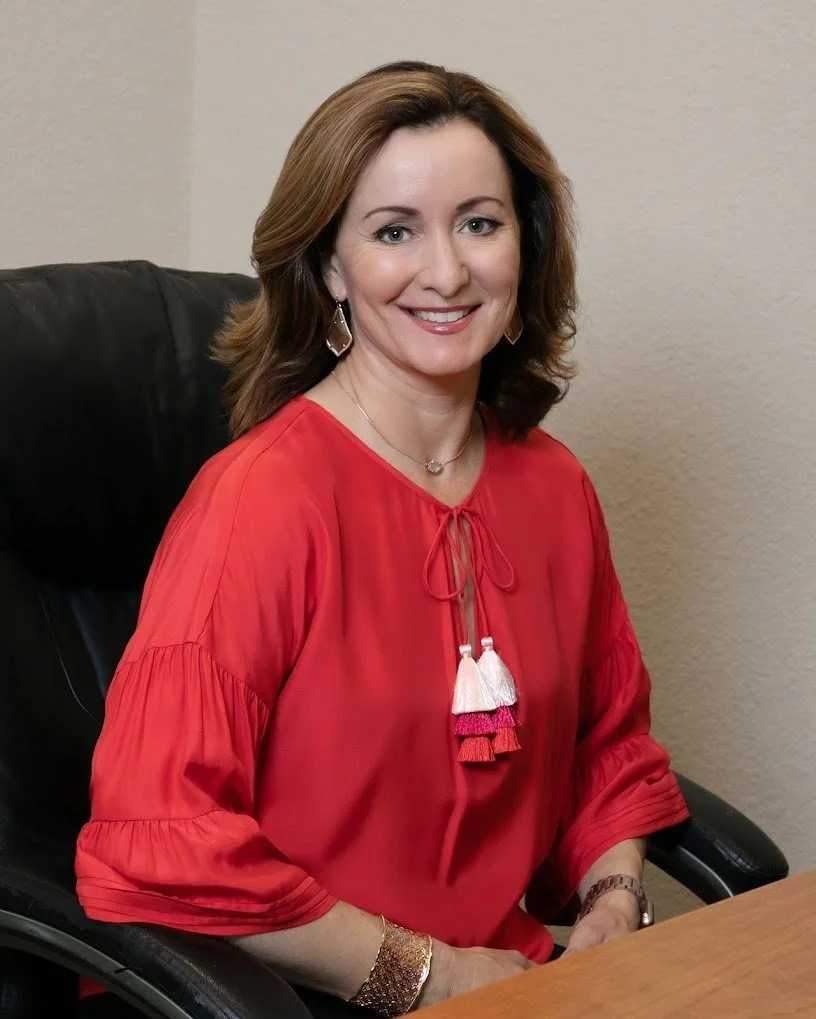 A woman with brown hair wearing a red blouse, earrings, a necklace, and a bracelet, sitting at a desk and smiling.