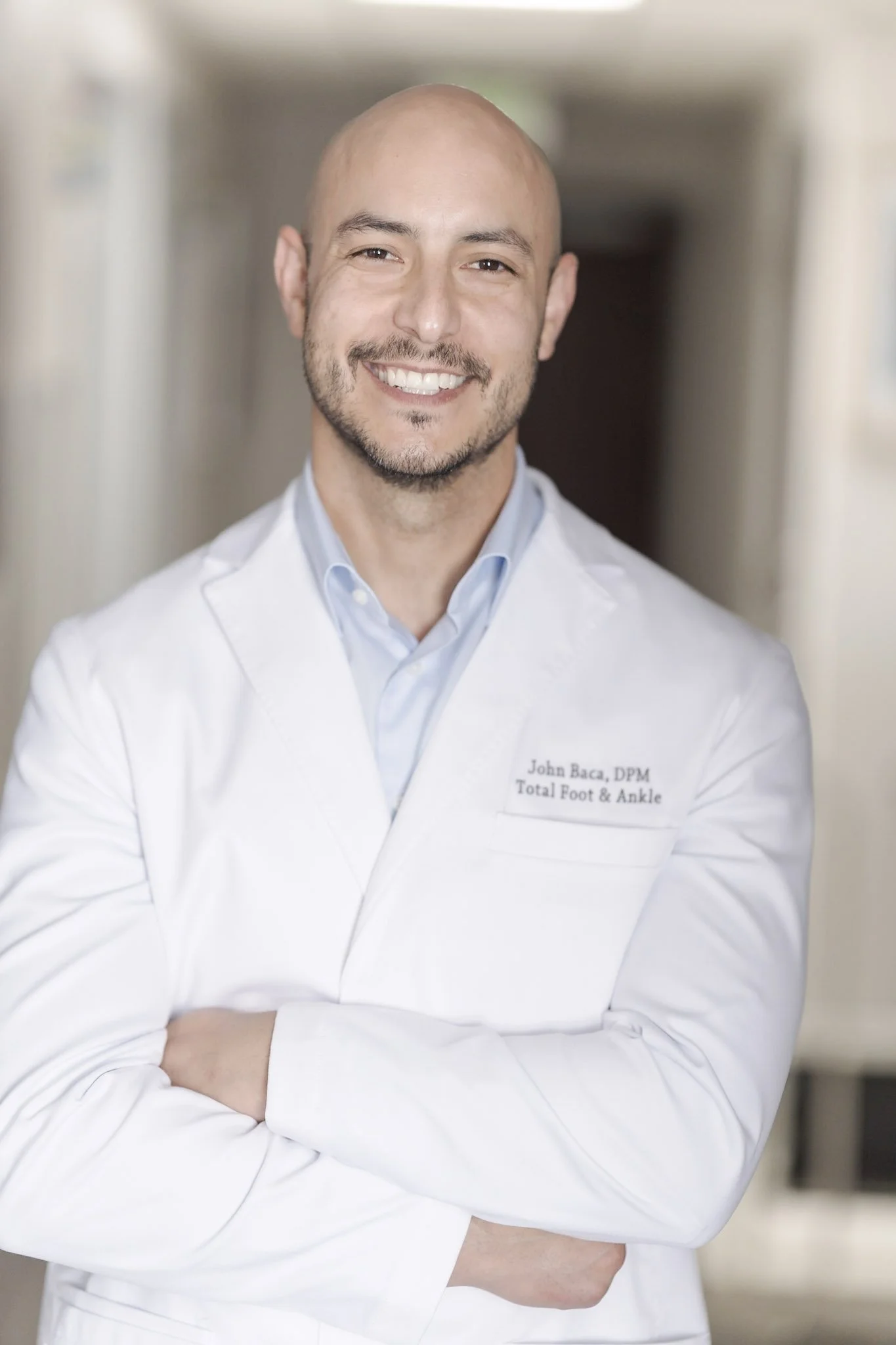 A smiling man with a bald head and beard standing in a hallway wearing a white medical coat with a name tag.