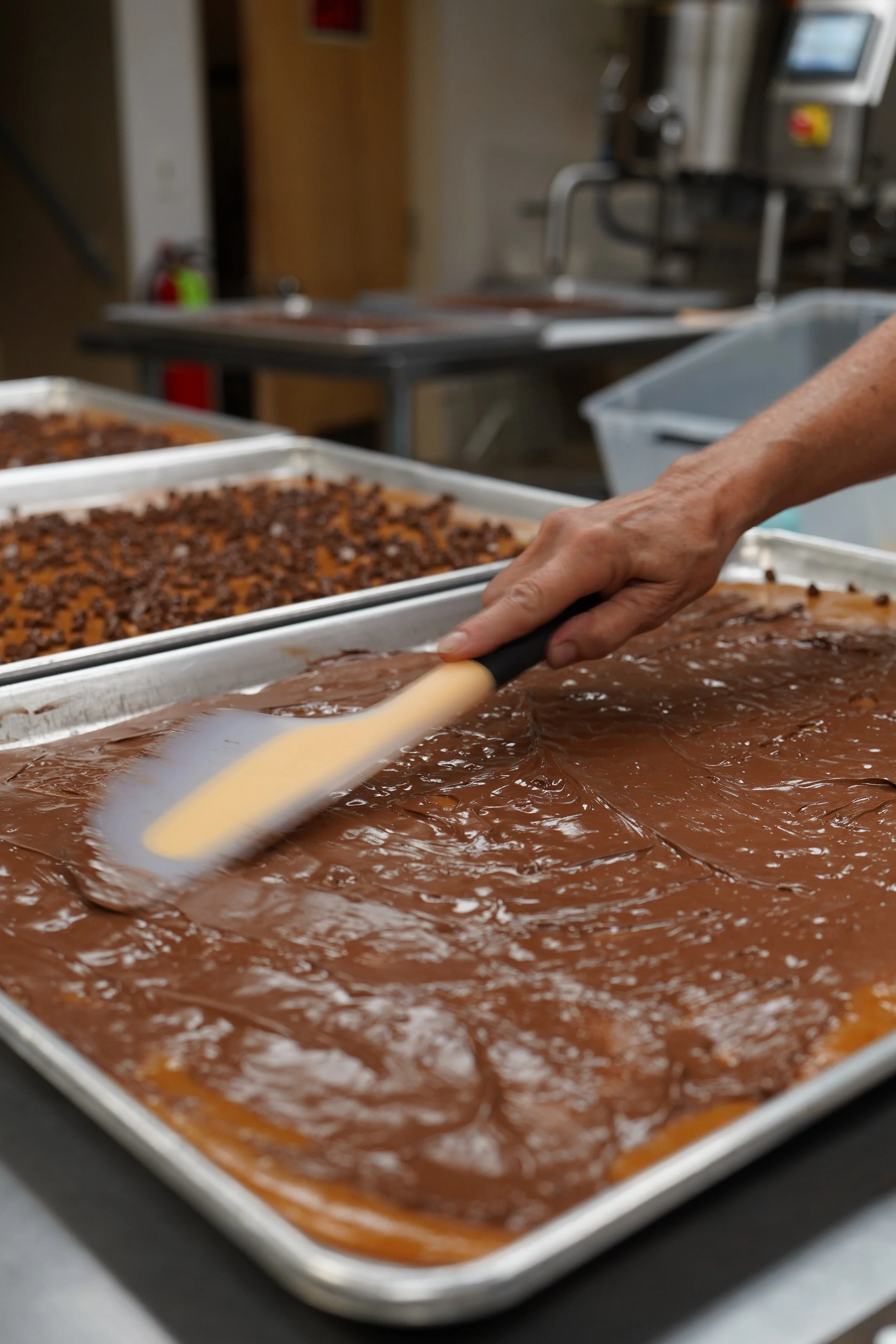 A person spreading chocolate frosting over a large baking sheet of brownie batter, with other trays of baked brownies in the background.