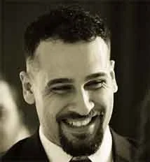 Close-up of a smiling man with dark curly hair and a beard, dressed in formal attire.