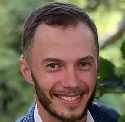A smiling man with short brown hair and a beard, wearing a blue checkered blazer and white shirt, standing outdoors with green foliage in the background.