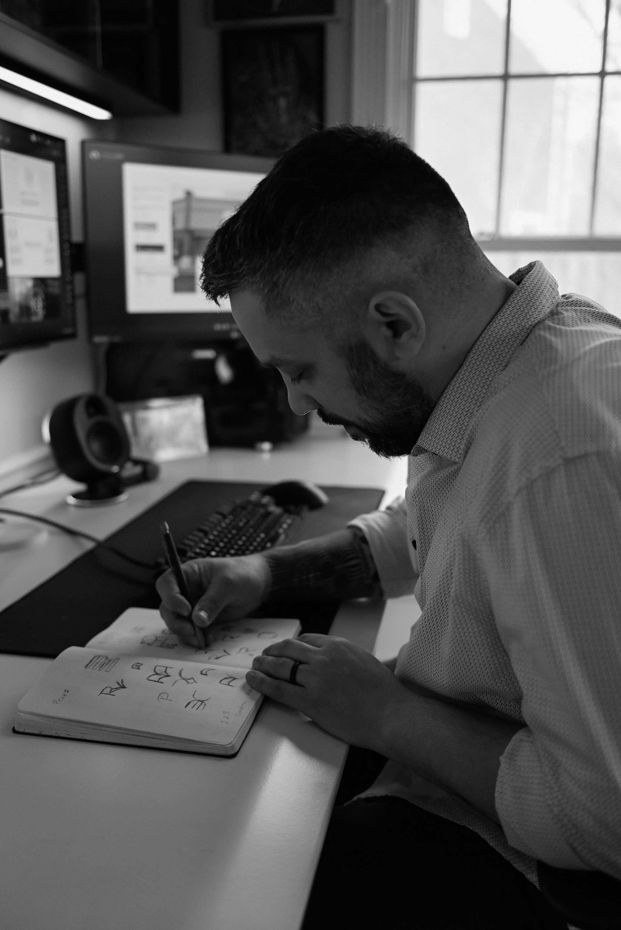 Clayton Hightower at his desk sketching. There are two computer monitors, a keyboard, and a speaker on the desk. The man appears to be practicing calligraphy or handwriting, working intently. The room has large windows allowing diffused.