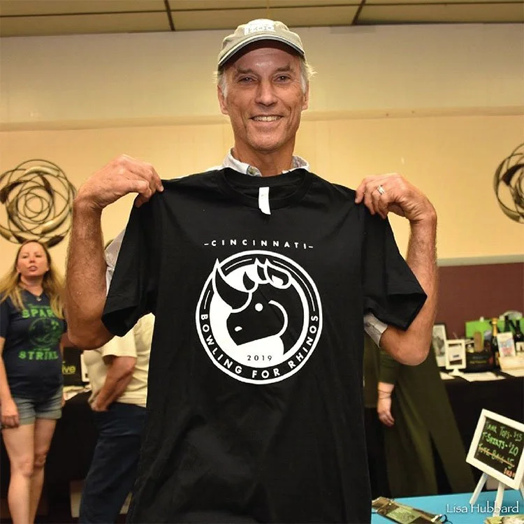 Thane Maynard smiling and holding up a black T-shirt with a unicorn logo and the text 'Cincinnati Bowling for Rhinos 2019' at an indoor event or fundraiser.
