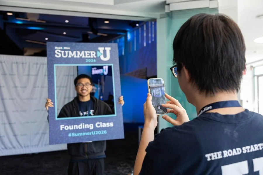 A student stand for a photo holding a Summer U frame board while another student takes his photo with their phone.