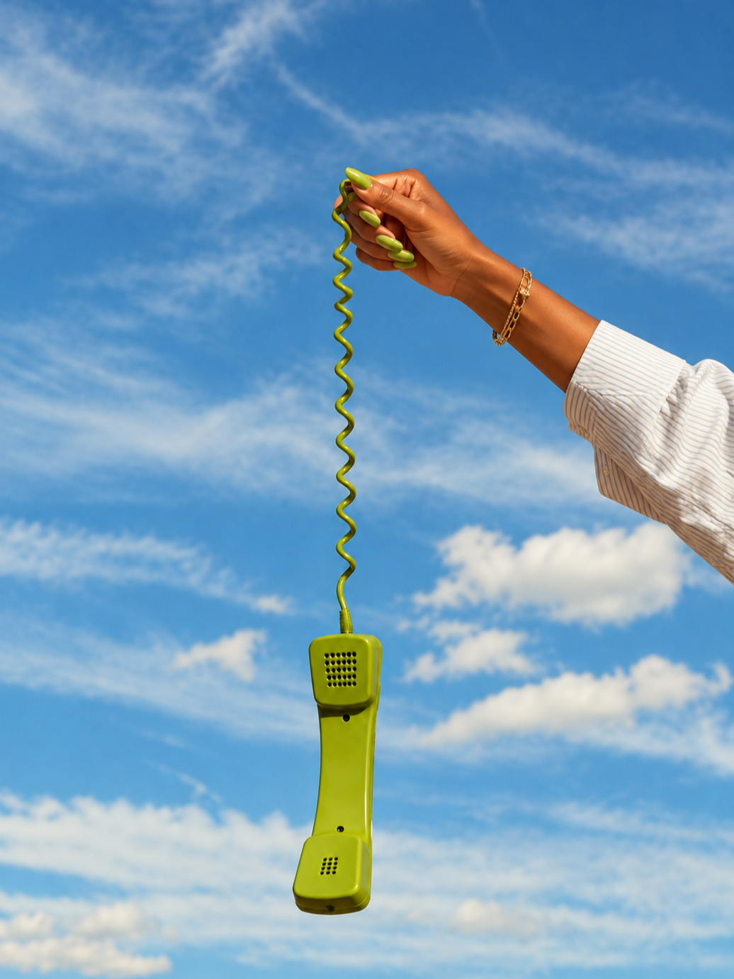 A person's hand with green nails and gold bracelet holding a green corded telephone receiver against a blue sky with clouds.