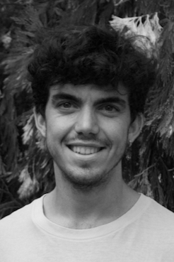 A young man with dark, curly hair, wearing a white t-shirt, smiling and standing outdoors in front of green leafy trees.