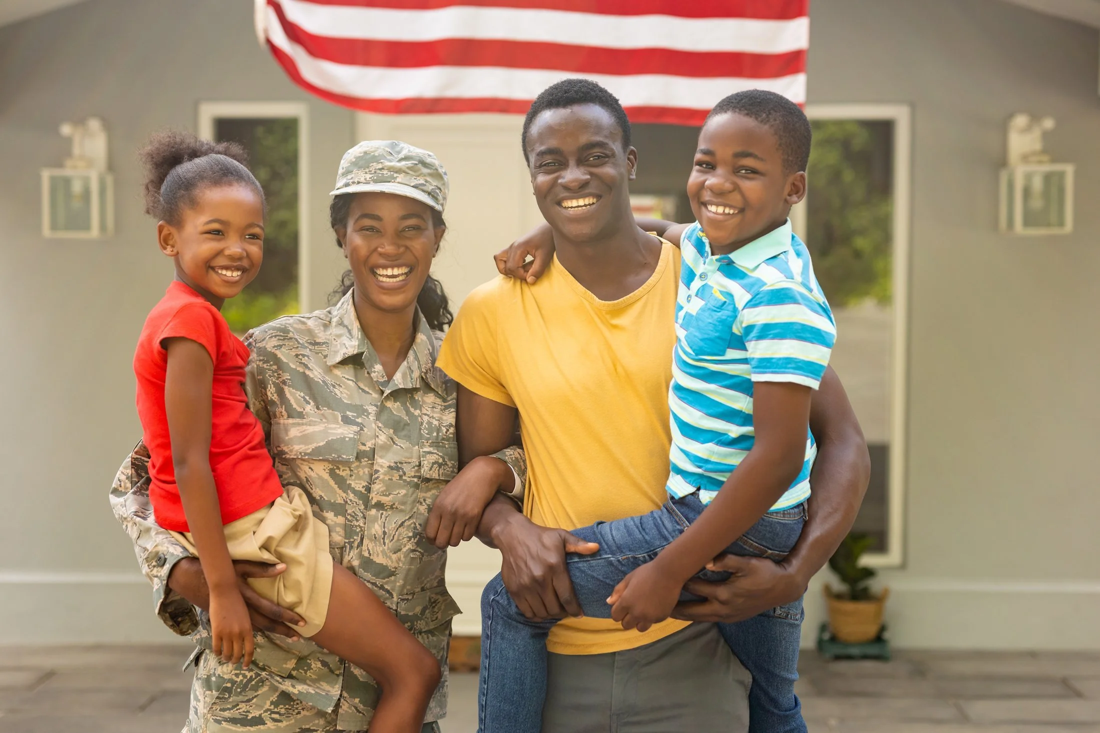 A smiling military family standing together in front of their home. They get to save an extra 10% while protecting their loved ones.