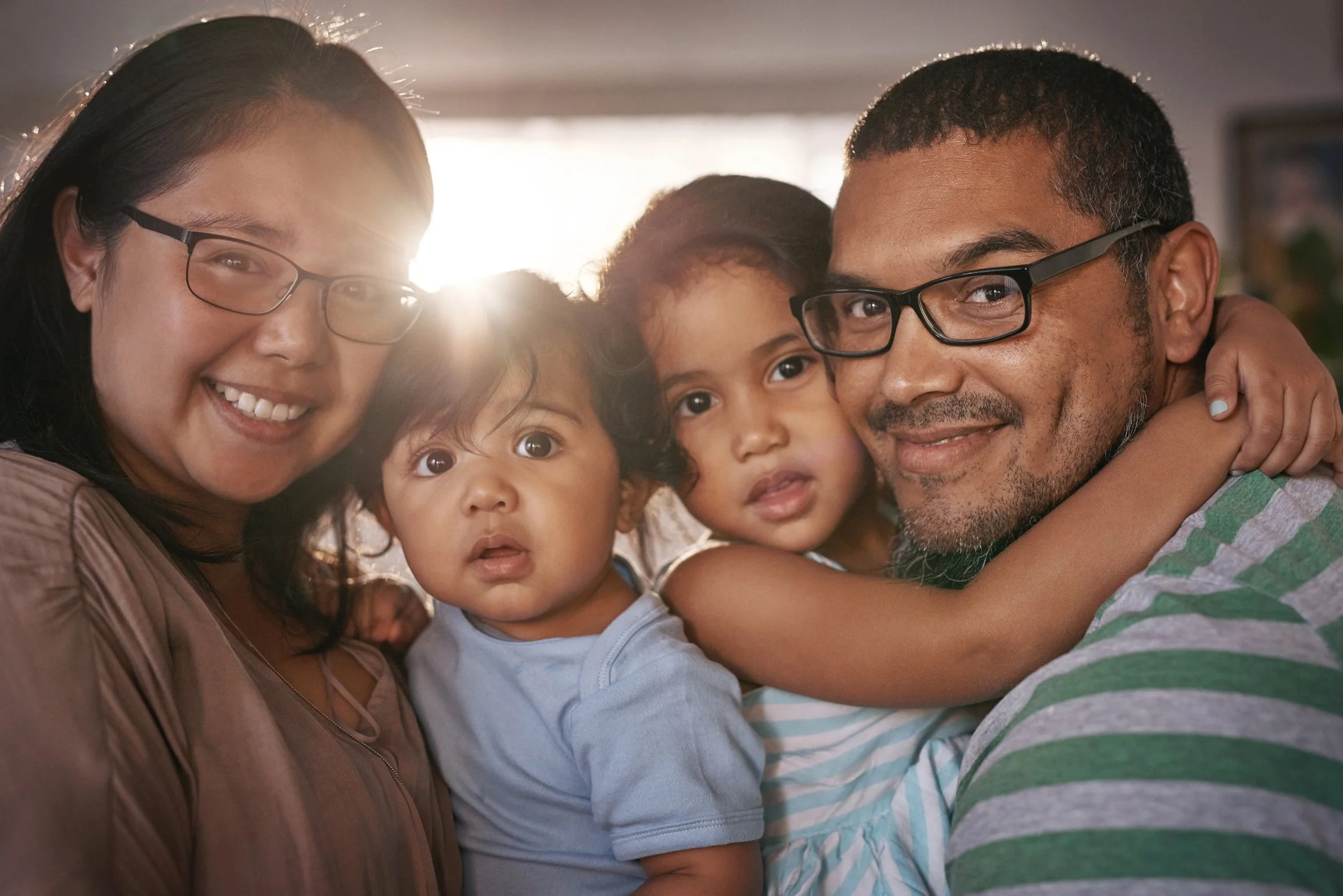 Parents holding two toddlers close, soft golden sun-flare behind them protected their family in case someone goes missing with Protect Our Family.