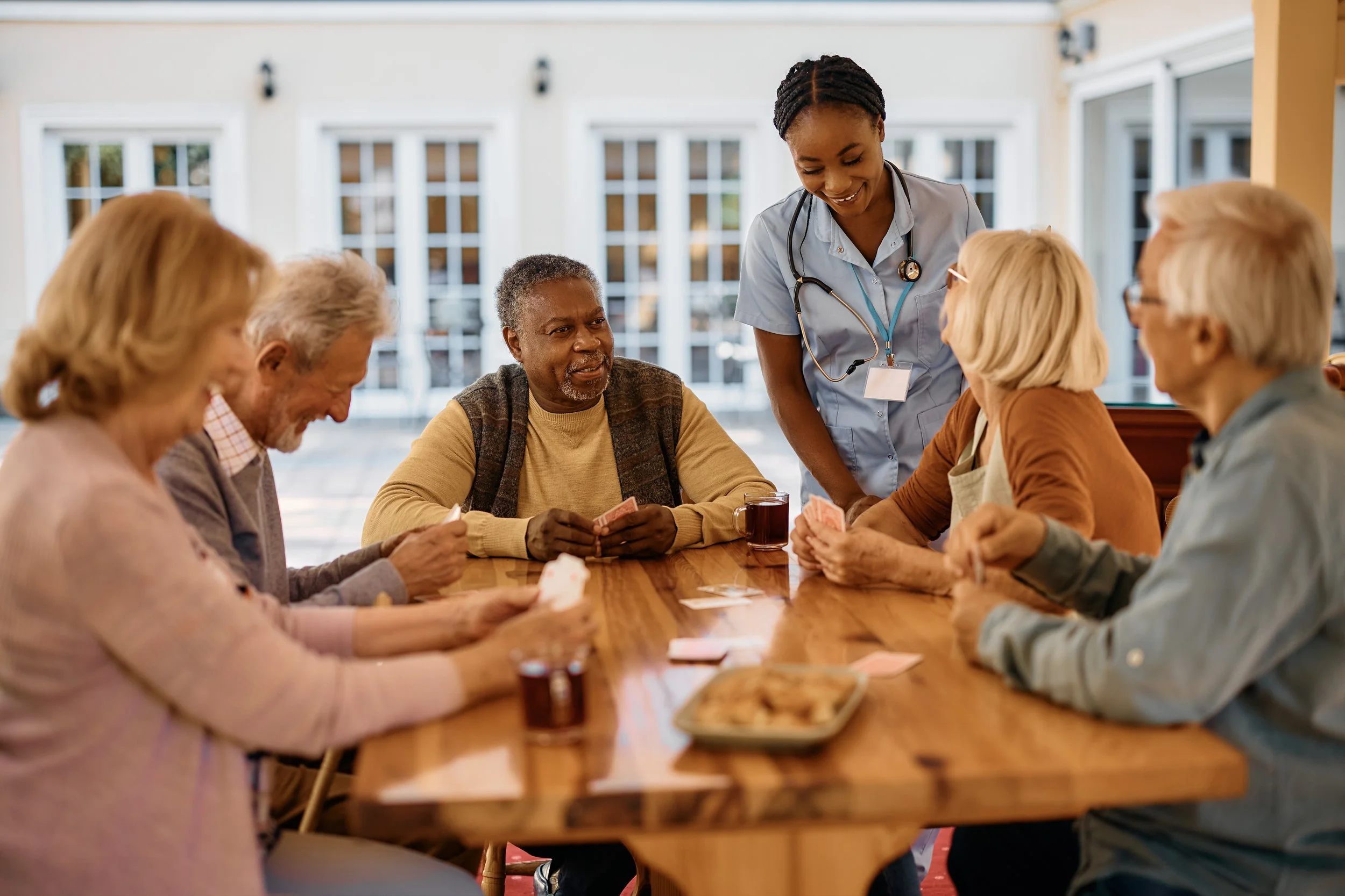 Seniors gather around a table to play a game with their caregiver. . Confident, calm presence because Protect Our Family is engaged.