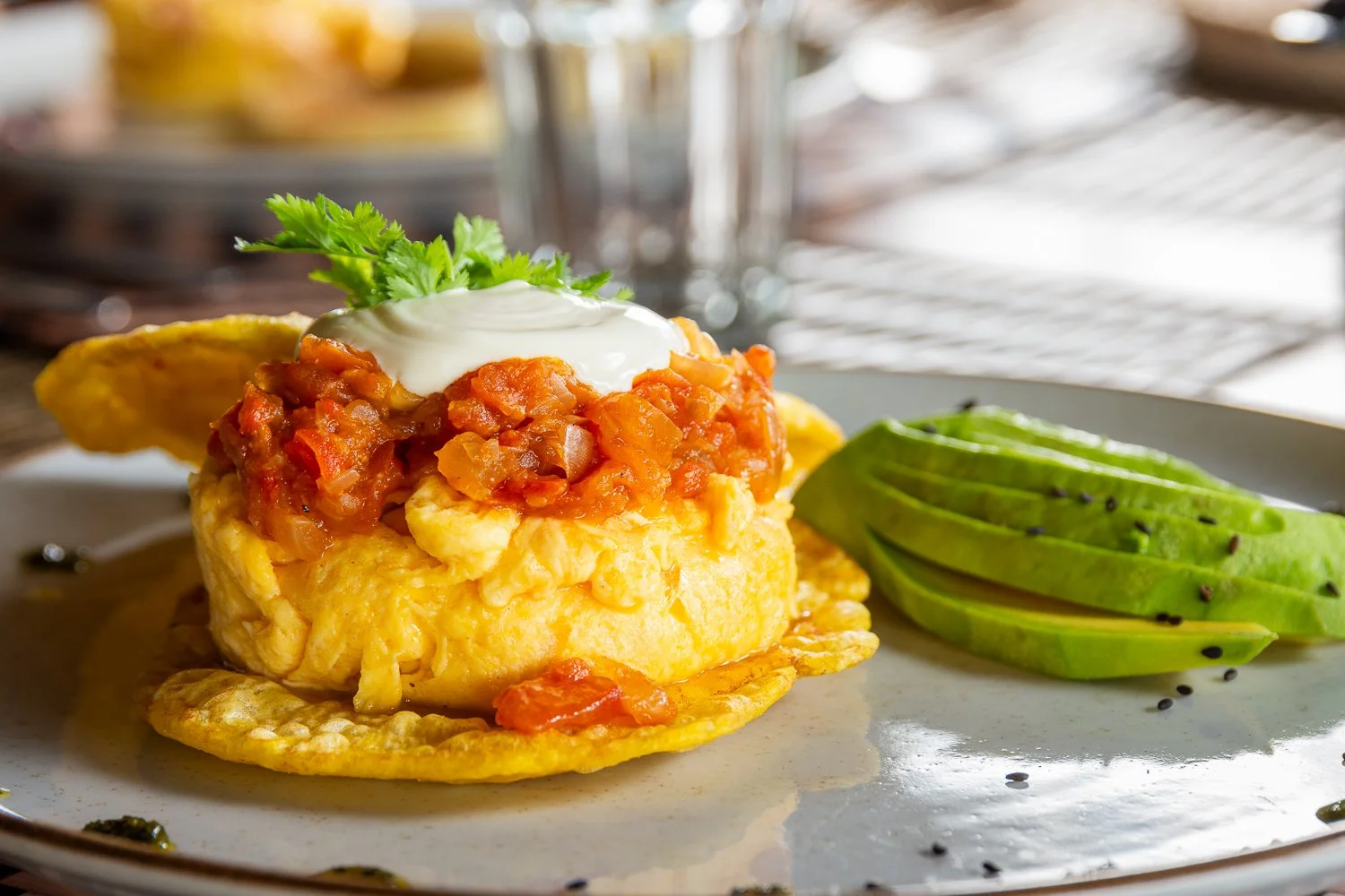 Person pouring orange sauce over a seafood dish with bananas and herbs on a white plate, set on a wooden table.