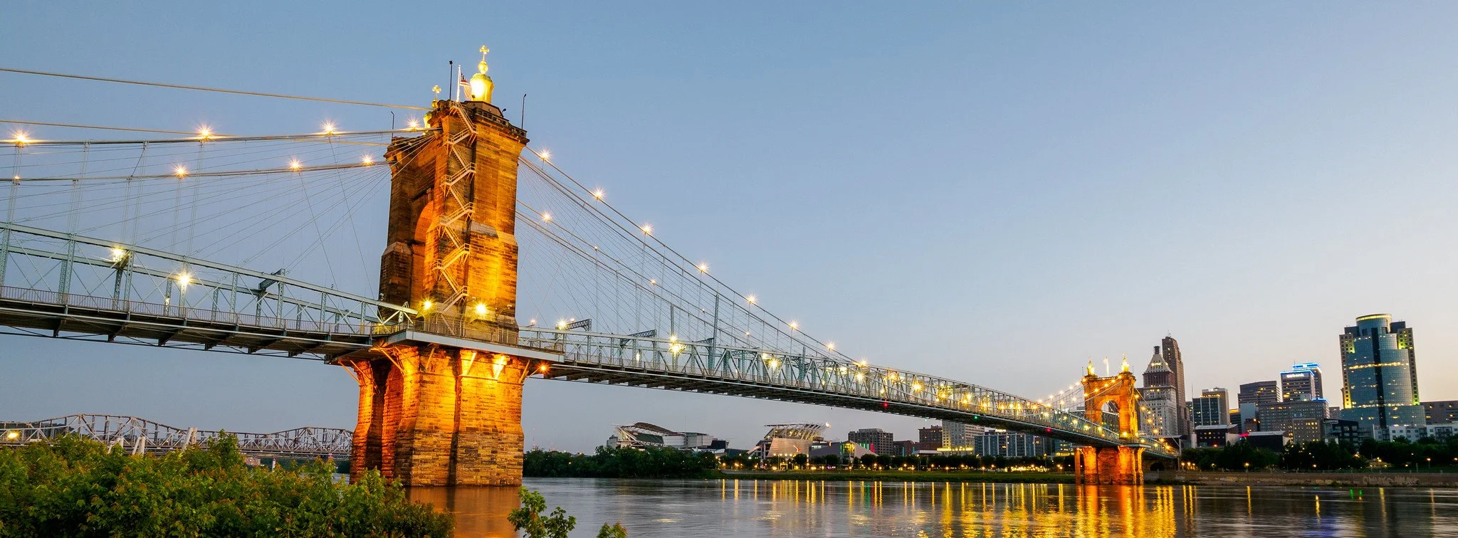 Night view of the John A. Roebling Suspension Bridge in Cincinnati, Ohio, with city skyline in the background and river in the foreground.