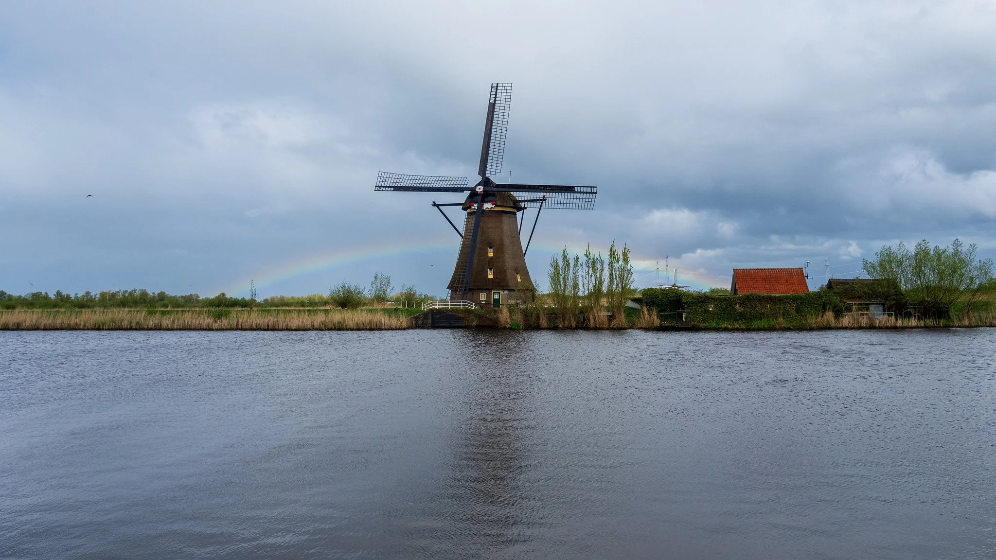 A traditional Dutch windmill next to a river with a rainbow in the cloudy sky.
