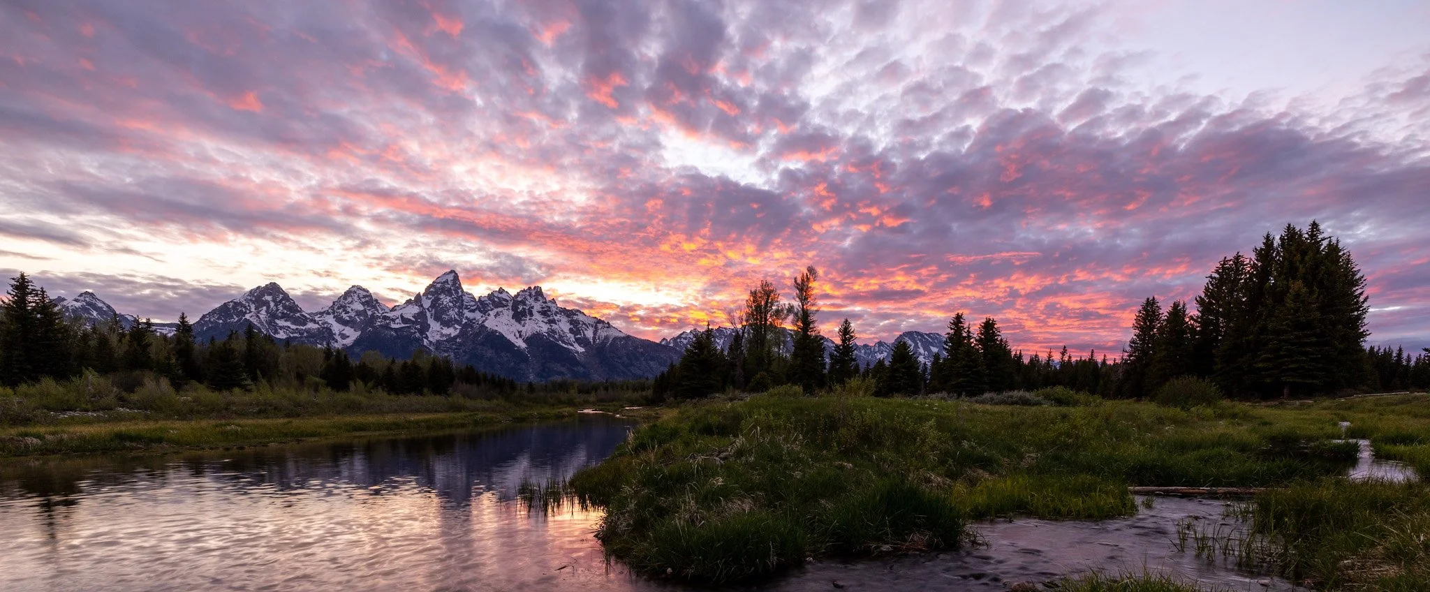 Sunset over snow-capped mountains, with colorful pink and purple clouds, trees, and a river in the foreground.