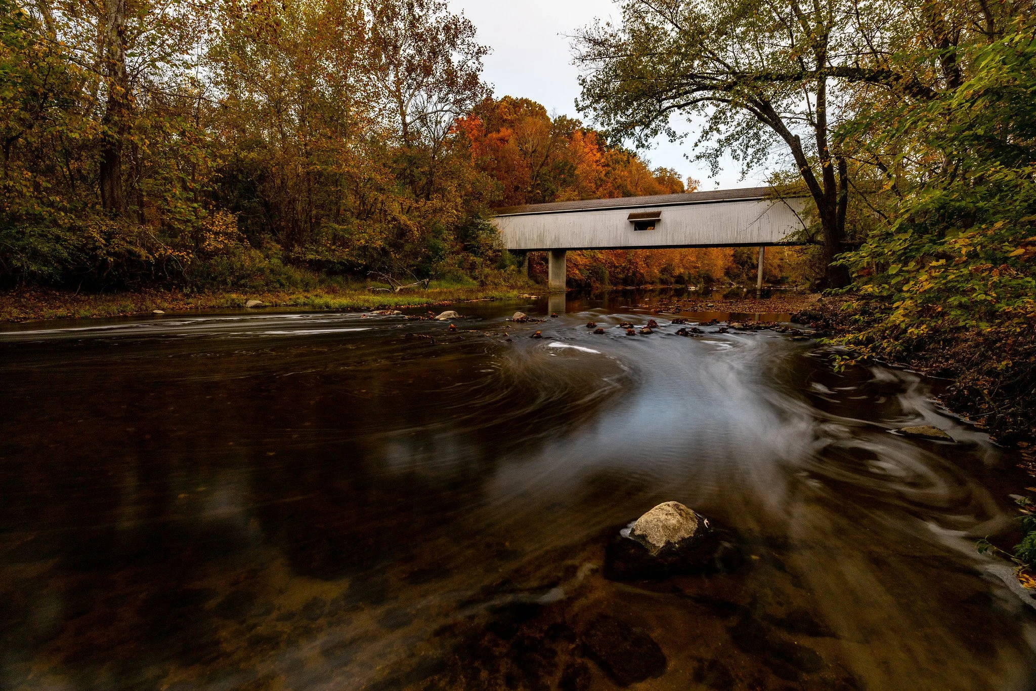 A river flowing under an old covered bridge surrounded by autumn trees with orange, yellow, and green leaves.