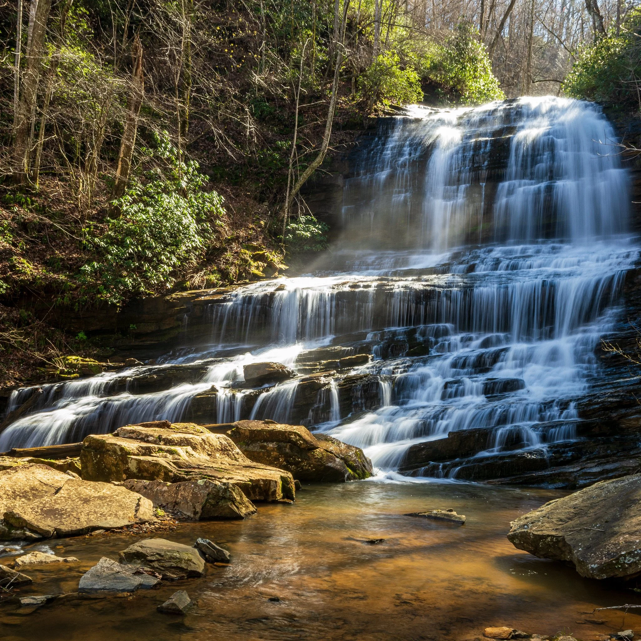 A multi-tiered waterfall flowing over rocks in a wooded area with trees and green bushes surrounding it.