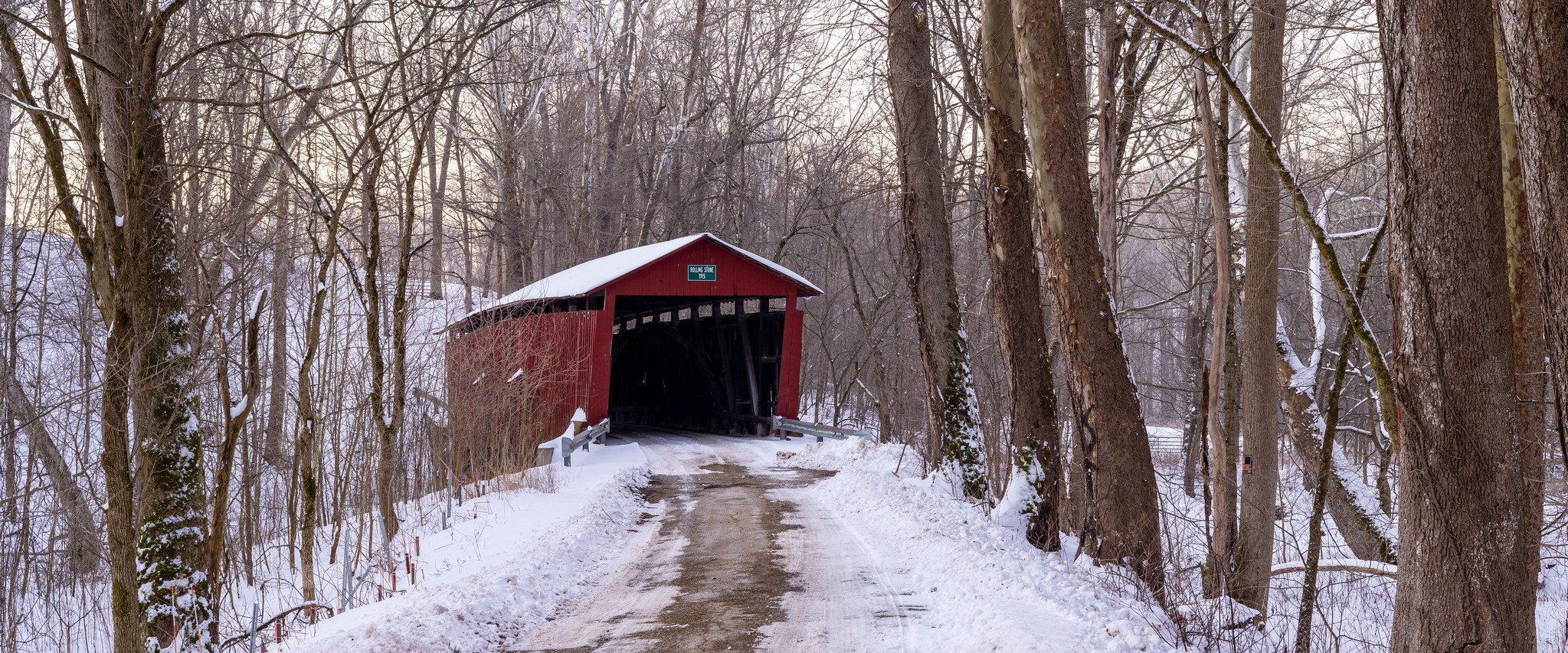 A snow-covered hiking trail leading to a red covered bridge in a forest during winter, with bare trees and snow on the ground.