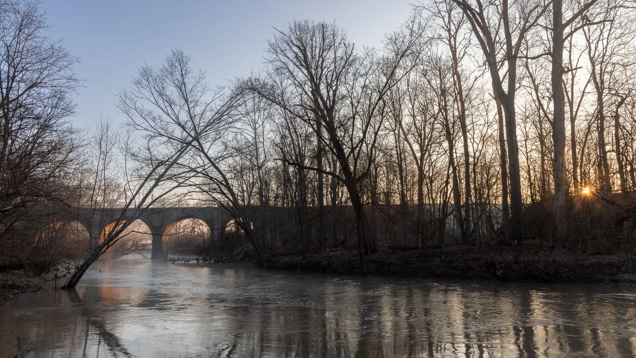 A river flowing through a leafless forest at sunset with a bridge in the background.